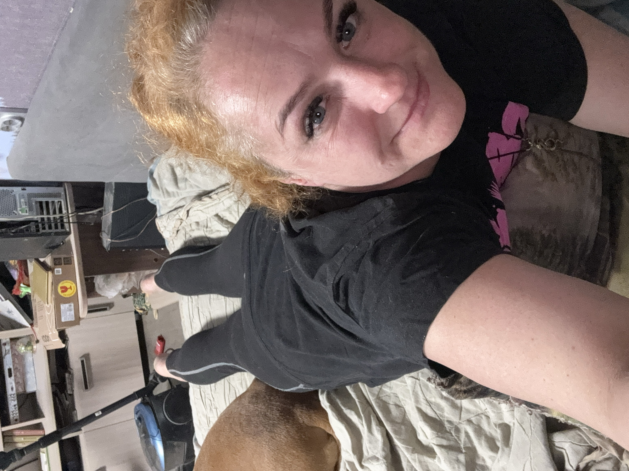 A person with fair skin and blonde hair is lying on a bed with a wrinkled white sheet. The person is wearing a black t-shirt with a pink design on the front and black leggings with grey stripes. A brown dog is lying near the person. To the left, there is a desk with a computer tower, boxes, and a vacuum cleaner. The location is in the city of Chaikovsky, Russia.