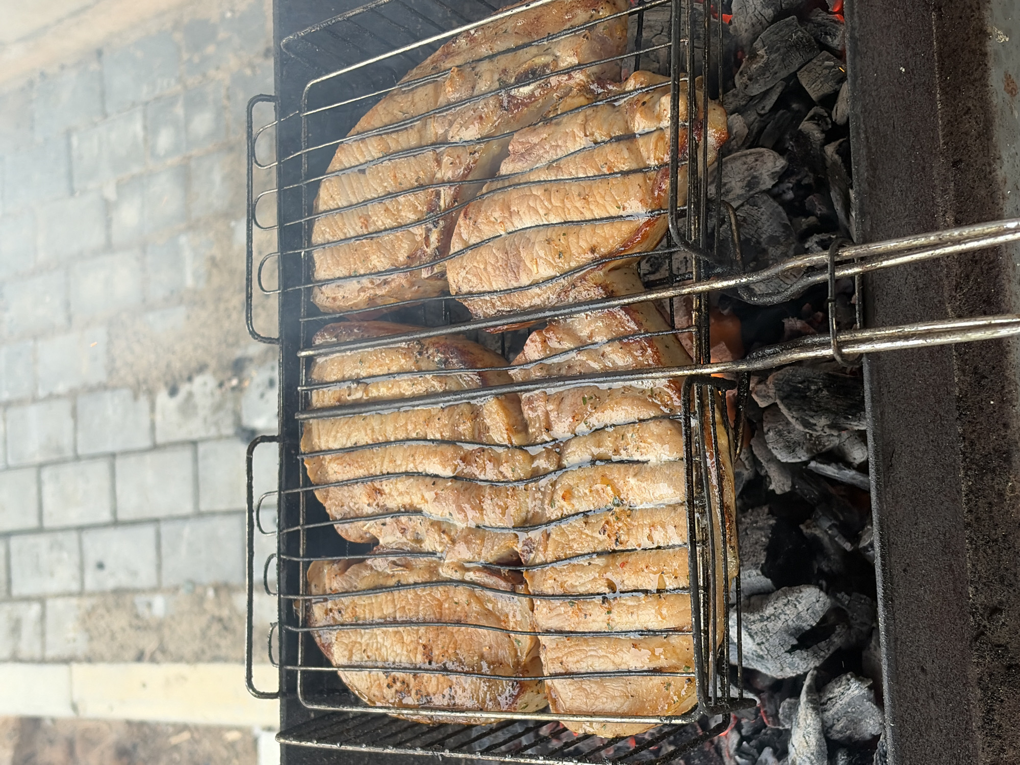 An overhead shot captures a grill basket filled with large pieces of meat, likely pork or beef steaks, cooking over hot charcoal in an outdoor setting. The meat, which appears to be seasoned with herbs and spices, is golden brown and glistening, suggesting it's actively searing and cooking. The grill basket holds multiple thick cuts of meat, arranged horizontally across its metal grates.
Below the meat, glowing red and grey embers of charcoal are visible, indicating a high heat source. The grill itself is a metal structure, possibly a mangal, with visible signs of use. Wisps of smoke rise from the grill, adding to the atmosphere of outdoor cooking.
In the blurred background to the left, light-colored brickwork or paving stones can be seen, hinting at a patio or yard area. The natural lighting suggests it is daytime. The scene evokes the enjoyment of preparing a meal outdoors. There is no visible text in the image. This activity is taking place in Chaikovsky, Russia.