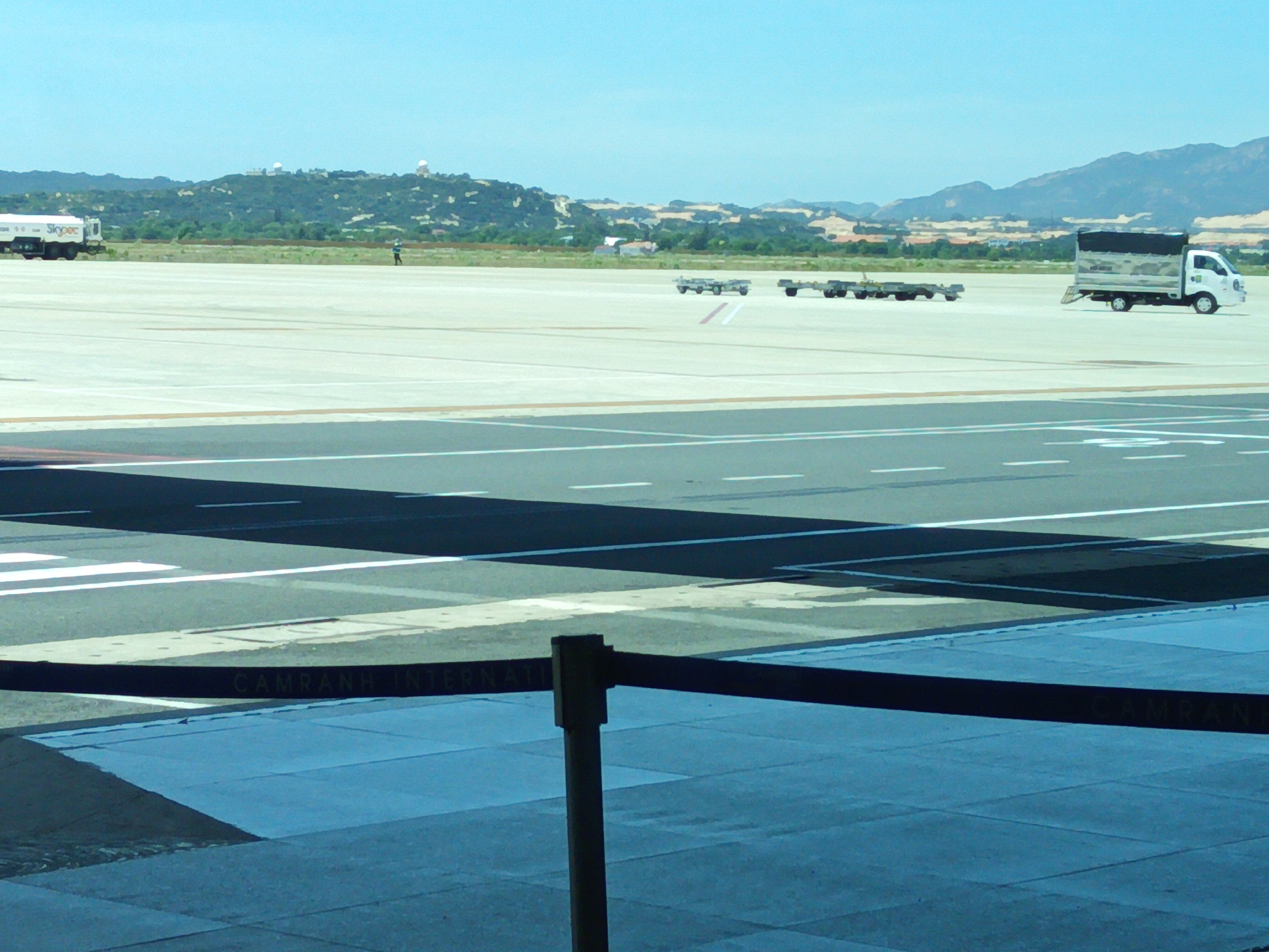 The image depicts an outdoor scene at an airport, likely within the vicinity of Bắc Cam Ranh, Vietnam, given the visible text "CAMRANH INTERN" on a dark barrier in the foreground. The foreground is dominated by a blue tiled area and a dark barrier with white text. Beyond this, a tarmac area is visible with various markings and shadows. In the mid-ground, several airport utility vehicles and equipment are present. To the left, a large white tanker truck with "Skypec" branding is parked. Further to the right, two sets of baggage carts or dollies are positioned on the tarmac, with another white truck with a covered bed visible in the far right mid-ground. A lone individual in a blue shirt is walking on the tarmac in the mid-ground, between the tanker truck and the baggage carts. The background features a landscape of rolling, tree-covered hills with some structures visible on the summits. The sky is clear and blue, suggesting a daytime setting. The overall impression is that of an active airport environment with ongoing ground operations.
