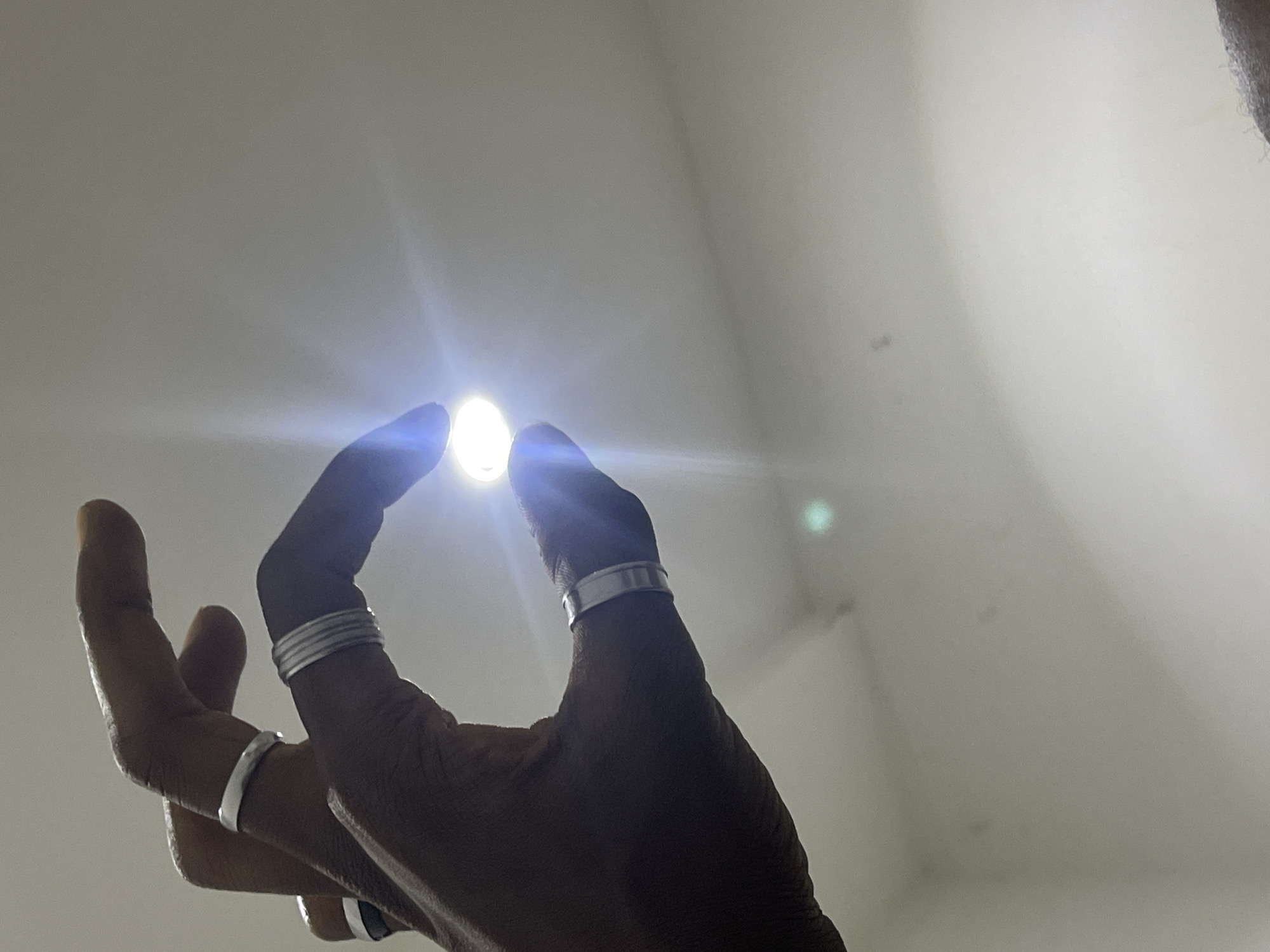 A close-up, low-angle shot captures a dark-skinned hand with several silver rings on its fingers, held against a plain, light-colored background. The hand is positioned as if pinching something between the thumb and index finger, though the object itself is not clearly visible. A bright light emanates from the area between the fingers, creating a lens flare effect and obscuring details. The background appears to be a ceiling or wall with subtle shadows and possibly faint discoloration, suggesting an indoor environment. The lighting conditions suggest the image was taken with a flash or a strong light source directed towards the camera. There are no other visible people or identifiable objects in the frame. The setting of Abuja, Nigeria, cannot be determined from the visual information present in the image.