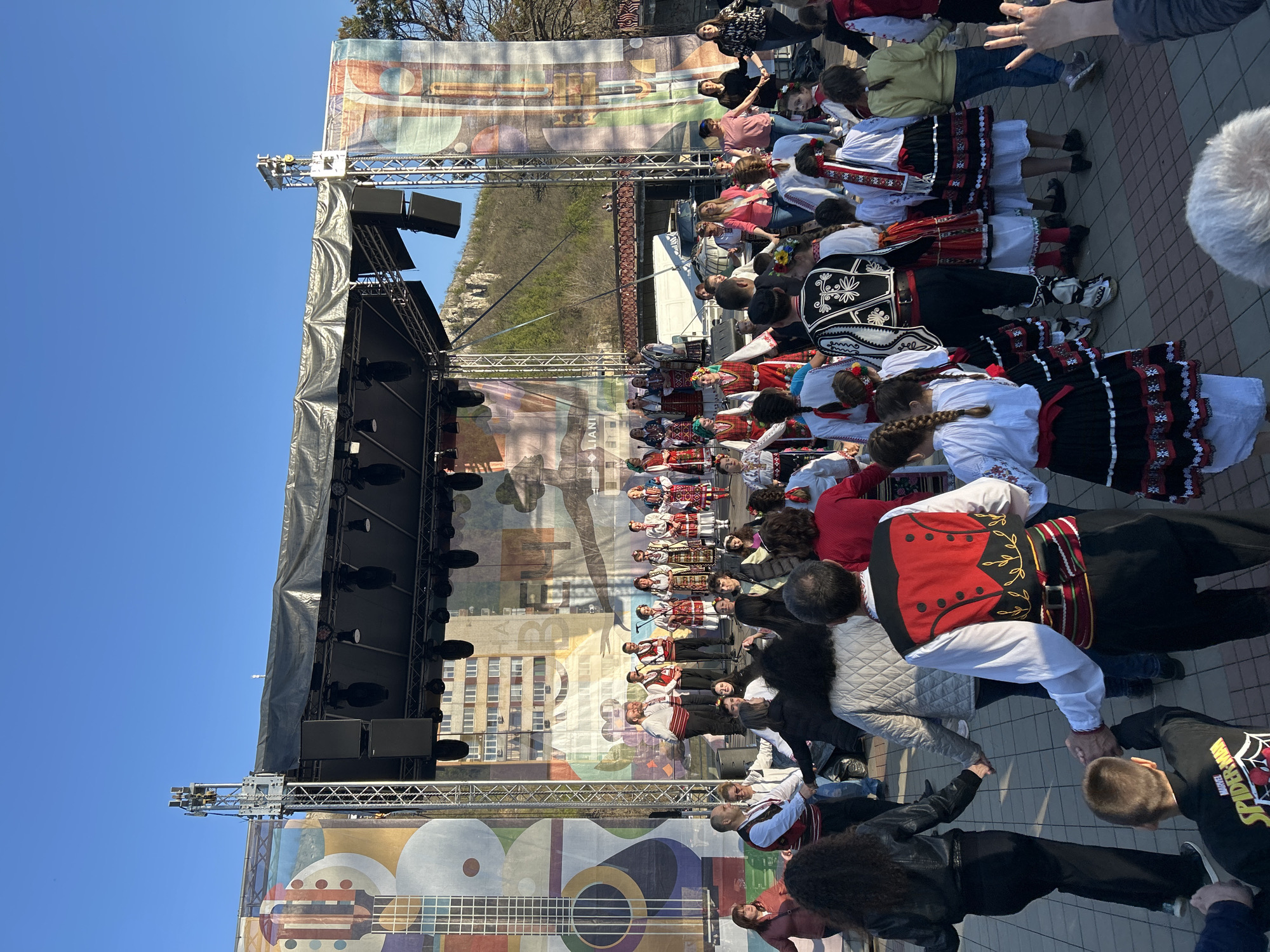 A group of people in traditional Bulgarian folk costumes are performing a dance in front of a stage in the city of Lovech, Bulgaria. The stage has a large backdrop featuring a guitar and other colorful designs. A small crowd is gathered, watching the performance. The sky is clear and blue. There are buildings in the background, including some high-rise apartments.