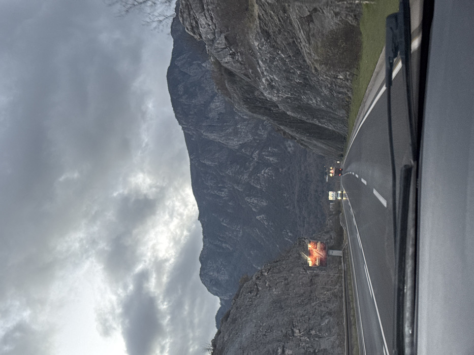 A road winds through a mountainous landscape under a cloudy sky. A large, gray mountain looms to the left, its rocky texture visible. The road, marked with white lines, leads towards two indistinct vehicles in the distance. Closer to the viewer, along the road, stand structures with glowing, possibly illuminated displays. The view is captured from inside a vehicle, with a portion of the dashboard or side visible on the right.