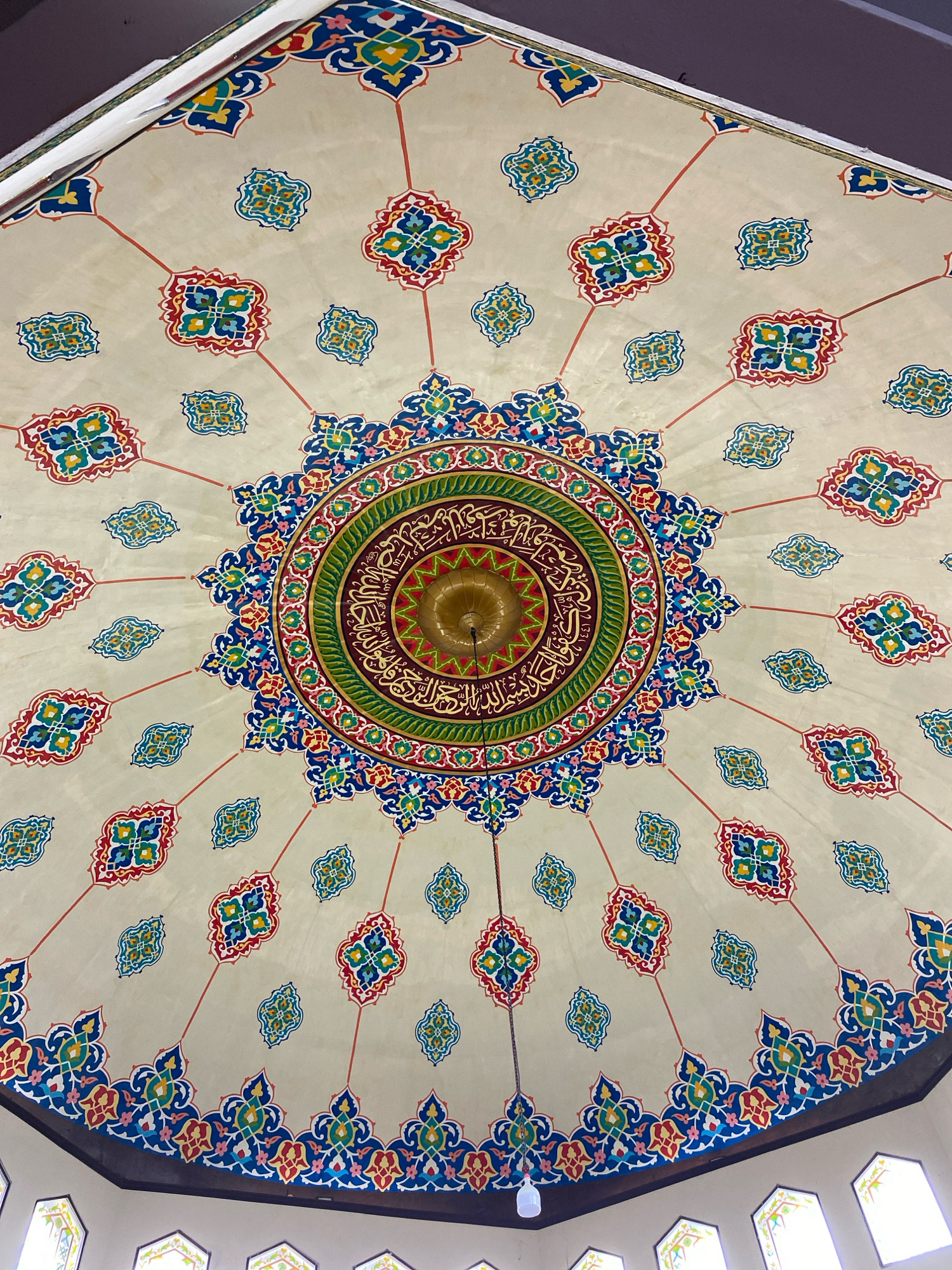 The image shows a detailed view of the ornate ceiling of the Islamic Center mosque in Lhokseumawe, Indonesia. The ceiling is a dome-shaped structure painted with intricate geometric patterns and calligraphy in vibrant colors such as red, blue, green, yellow, and gold.
At the center of the dome, a large, circular medallion features elaborate Islamic calligraphy in gold, surrounded by concentric rings of colorful floral and geometric motifs. Radiating outwards from this central medallion are lines of red paint, connecting to smaller, repeating decorative elements scattered across the ceiling. These elements are typically square or diamond-shaped, filled with a similar palette of colors and intricate designs. The dome's edge is adorned with a band of decorative patterns that transition into the surrounding architectural features.
A golden chandelier hangs from the very center of the dome, its intricate design echoing the patterns on the ceiling. Towards the bottom of the image, the edge of the dome meets the upper portion of the walls, where windows with decorative stained glass are partially visible. A white light fixture hangs from the ceiling, partially obscuring the view of the lower part of the central medallion and the chandelier. The overall impression is one of rich artistic craftsmanship and religious devotion, characteristic of Islamic architecture.