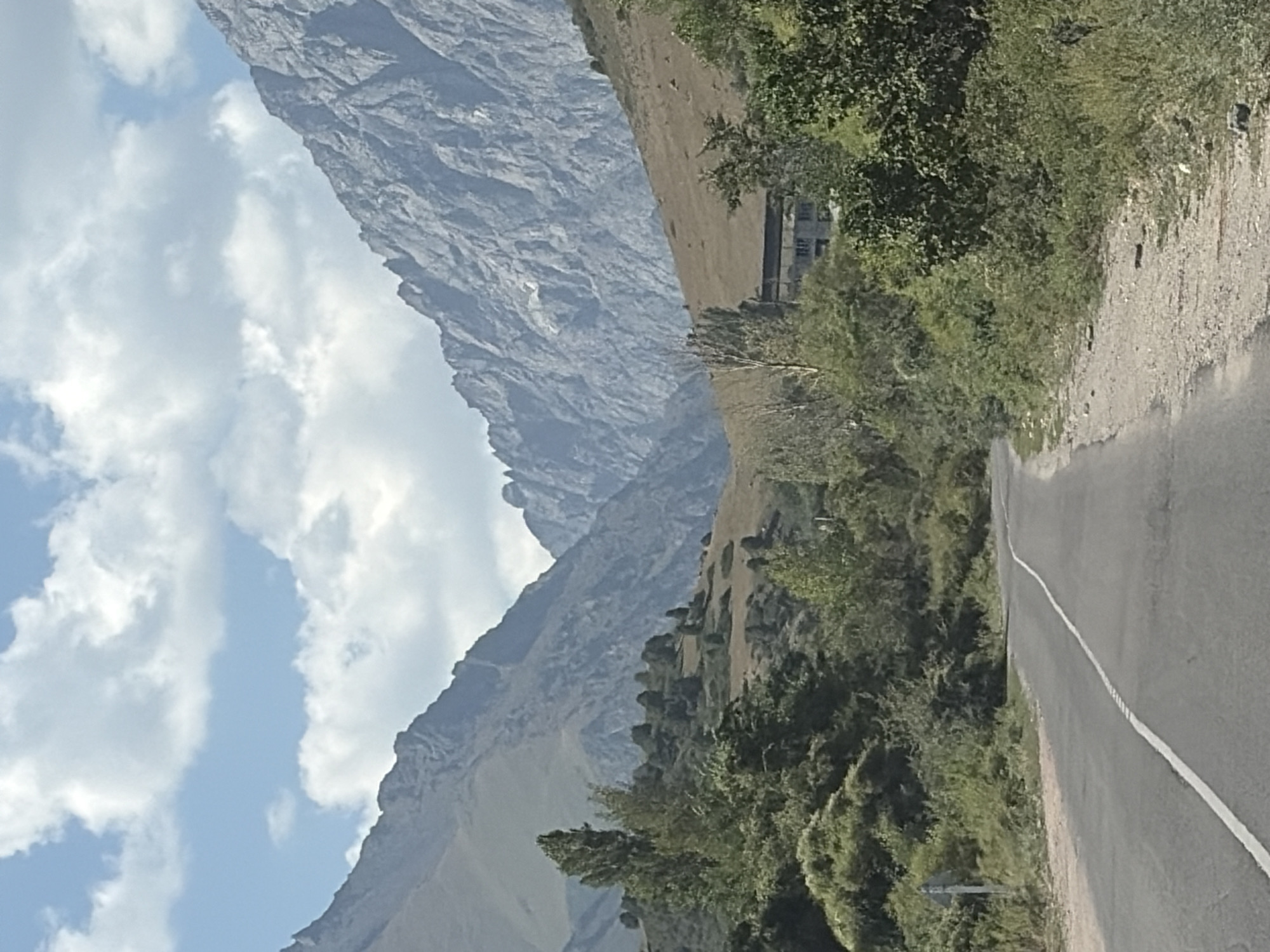 A paved road curves to the left, bordered by sparse green vegetation and trees. In the background, rolling hills covered in dry grass rise towards imposing, rugged mountains under a partly cloudy sky. A building with a dark roof and visible windows is partially obscured by trees on the right side of the frame.