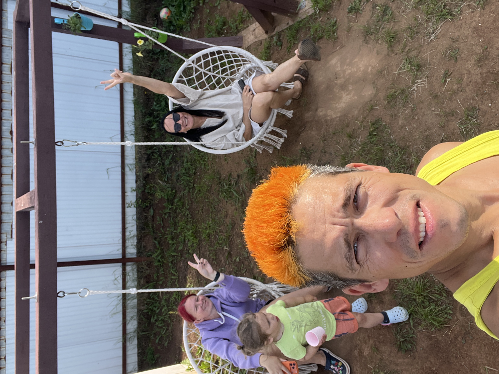 The image is a selfie taken outdoors, likely in a backyard, with a swing set structure in the background. In the foreground, a man with bright orange hair is smiling at the camera. To his left, a young child wearing a green shirt and orange shorts is standing, holding a pink cup. Behind the child, a woman with red hair is sitting in a white hanging chair, making a peace sign. Further back, another woman wearing sunglasses and a light-colored top is seated in a similar white hanging chair, also making a peace sign. The environment appears to be a grassy area with dirt paths, and a white fence is visible behind the swing set. The scene is set during daylight. The location is confirmed as Chaikovsky, Russia.