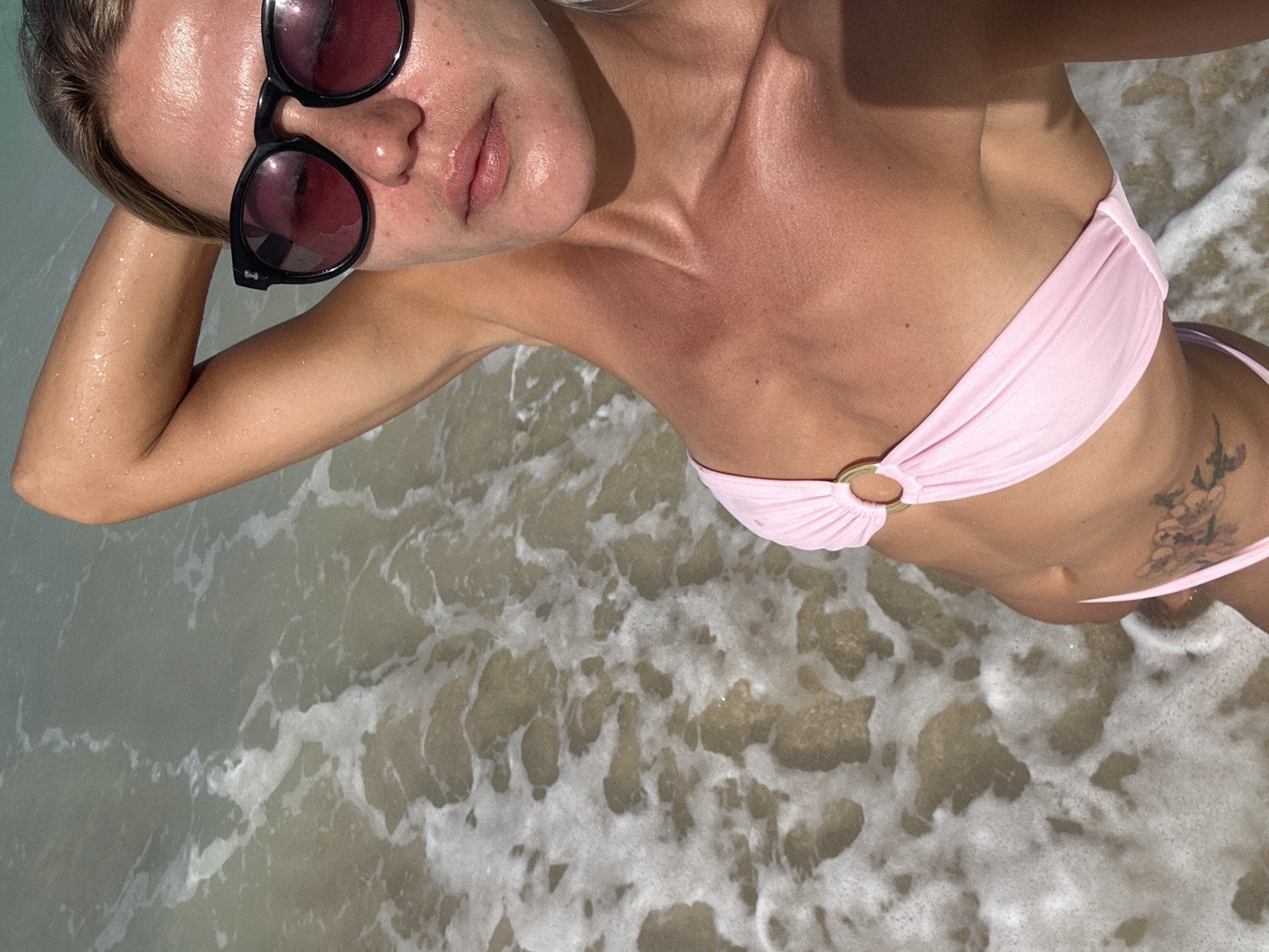 A woman is posing for a selfie at the beach, with the ocean in the background. She is wearing a pink bikini and sunglasses. Her hair is blonde and pulled back. The water is clear and shallow, with some waves and foam. The woman appears to be in good health and is enjoying her time at the beach. The location is Kuta Selatan, Indonesia.