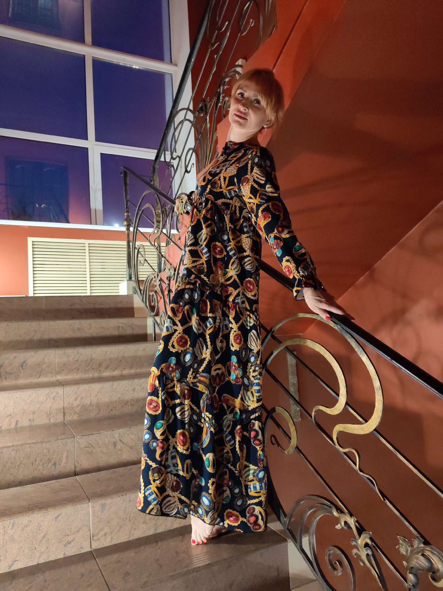 A woman with short, blonde hair and fair skin is standing on a staircase. She is wearing a long-sleeved, floor-length dress with a dark background and a colorful pattern of chains, jewels, and shells. Her left hand is resting on the ornate metal railing of the staircase. The staircase has brown, textured steps and a reddish-orange wall behind the railing. A large window with multiple panes is visible in the background, reflecting a dim, purplish light, suggesting it is nighttime or indoors with artificial lighting. There is a vent below the window. The woman is barefoot. She appears to be looking towards the camera with a neutral expression. The scene suggests an indoor setting, possibly a building with decorative architecture.