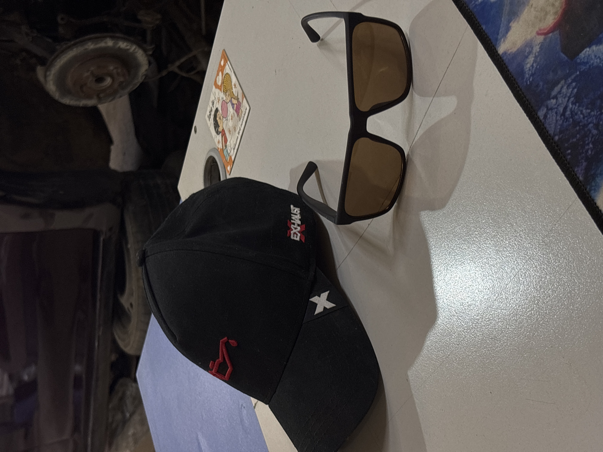 A black baseball cap, a pair of sunglasses, and a sticker are placed on a white surface. The cap has red embroidery. The sunglasses have brown lenses. A dark, indistinguishable area is visible behind the objects. This image was not taken in Chaikovsky, Russia.