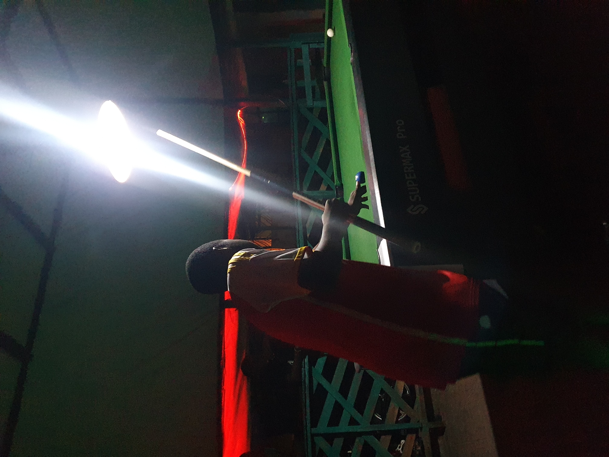 In Lokoja, Nigeria, a young Black male, seen from behind, is playing billiards under a bright overhead light. He wears a red and white shirt. He is positioned to strike a blue cue ball with a cue stick, aiming at a white ball on the green felt of a Supermax Pro pool table. The environment appears to be an indoor recreational space, possibly a bar or club, with a dimly lit background featuring red accent lighting and a green railing.