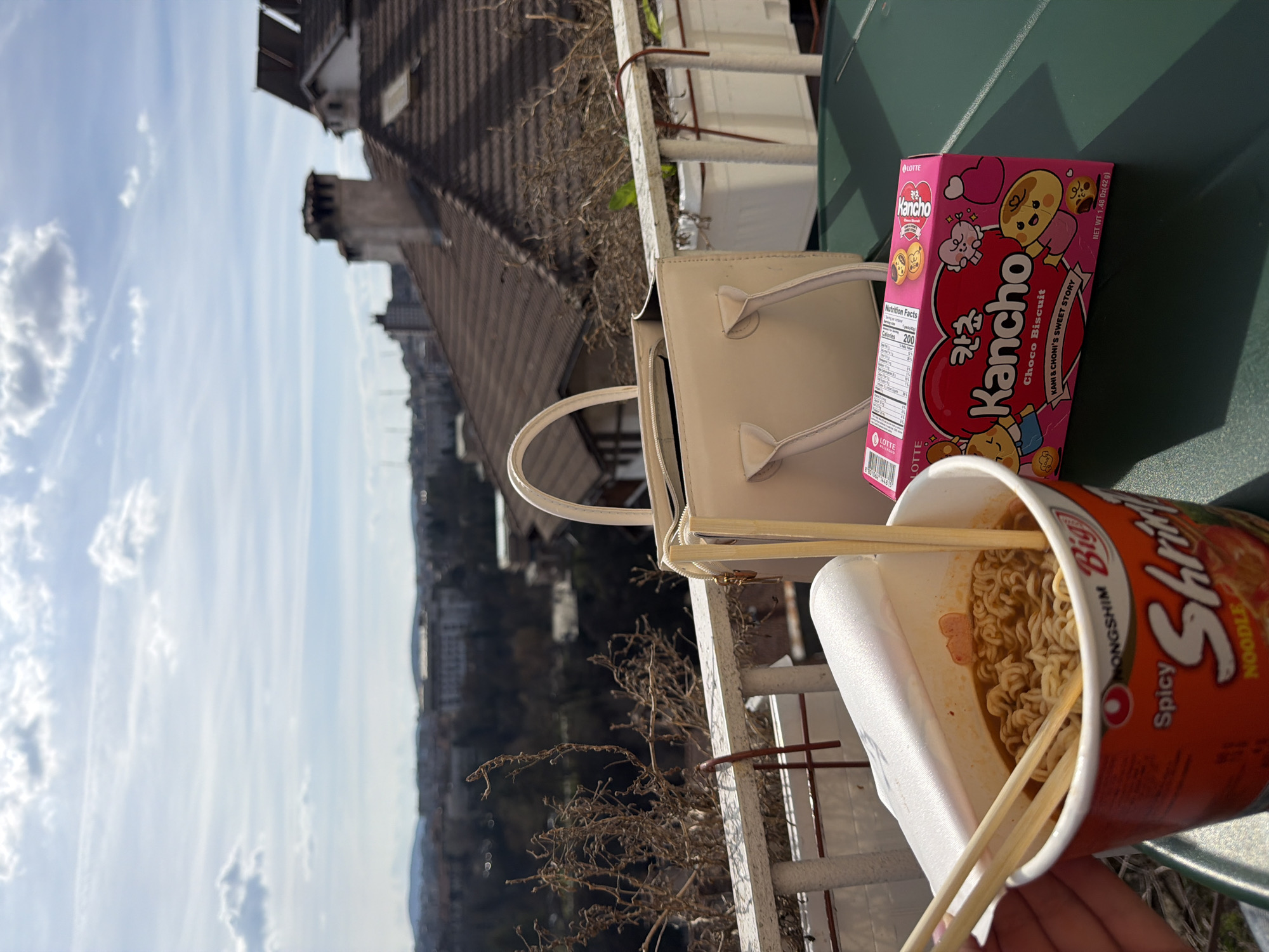 The image depicts a balcony or terrace overlooking the city of Veliko Tarnovo, Bulgaria, on a clear, sunny day with scattered clouds. In the immediate foreground, a person's hand with light skin tone holds a red and white Nongshim Big Spicy Shrimp Noodle instant ramen cup, open to reveal cooked noodles in a reddish broth with several small pieces. Wooden chopsticks are inserted into the noodles. To the right, on a dark green table surface, rests a pink box of Lotte Kancho Choco Biscuit, featuring cartoon characters. Behind the biscuit box, an open beige handbag with two top handles stands upright.
The midground features a white metal railing, likely part of the balcony, with several white plant pots attached. These pots contain dry, bare branches of a plant. Beyond the railing, the cityscape of Veliko Tarnovo unfolds. Closest are tiled rooftops of buildings, predominantly brown, featuring multiple chimneys and some dormer windows. Further in the background, a dense urban environment with various buildings, including a larger, lighter-colored structure, stretches towards distant, partially tree-covered hills. The trees closer to the foreground and within the city appear largely bare, suggesting a late winter or early spring season. Natural sunlight illuminates the scene, creating distinct shadows from the objects.