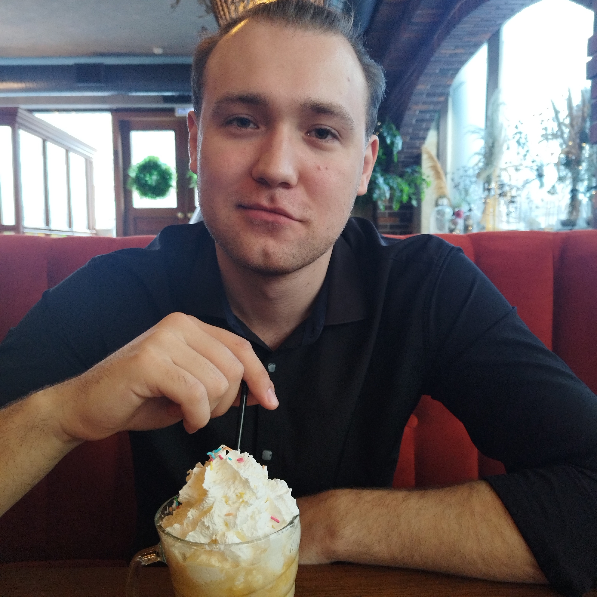 A young man, likely in his 20s, with light brown hair and light stubble, looks directly at the camera with a neutral expression. He is seated at a wooden table in what appears to be a cafe or restaurant in Yekaterinburg, Russia, identifiable by the red upholstered booth he occupies and the surrounding decor. He is wearing a dark, possibly black, collared shirt.
In front of him, on the table, is a clear glass mug holding a light-colored beverage, generously topped with whipped cream and colorful sprinkles. He holds a black straw in the drink with his right hand. His left forearm rests on the table.
The background reveals a well-lit interior with large windows. To his left, a wooden door with a green decorative wreath is visible through a windowed partition. To his right, an arched brick structure frames another large window, allowing bright daylight into the space. Various decorative elements, including plants and what appear to be festive or dried arrangements, are seen near the windows. The overall ambiance suggests a comfortable, daytime setting. There is no visible text in the image.