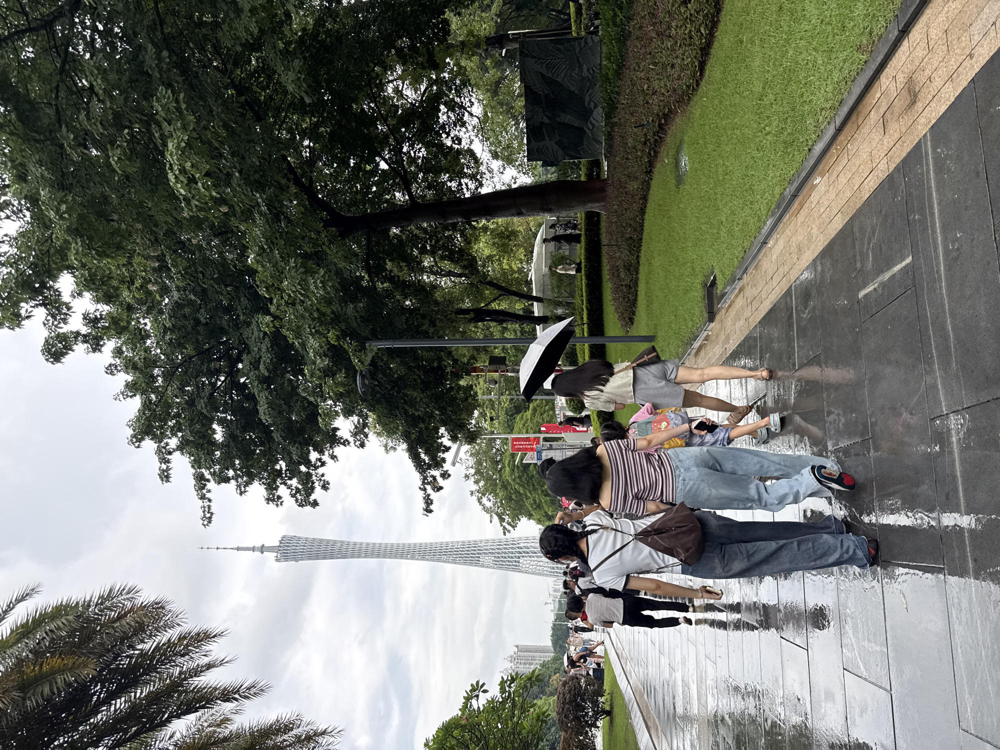 The image depicts a paved walkway in a park with lush green grass and trees. In the background, the Canton Tower, a prominent landmark in Guangzhou, China, rises against a cloudy sky. Several people are walking on the wet pavement, some under umbrellas, indicating it might be raining or has recently rained. The focus is on a group of four people walking away from the camera. The person in the foreground on the left has dark hair in braids and wears a white t-shirt and a brown bag. To their right is another person wearing a striped off-the-shoulder top and jeans. A child in a pink shirt and backpack walks between them. The fourth person, on the right, wears a light-colored top and grey shorts, holding a black umbrella. The walkway is made of large dark stone slabs with reflections from the wet surface. There are also planters with dark red and green foliage along the edge of the grass. The overall scene suggests a public park area in the Tianhe District of Guangzhou.