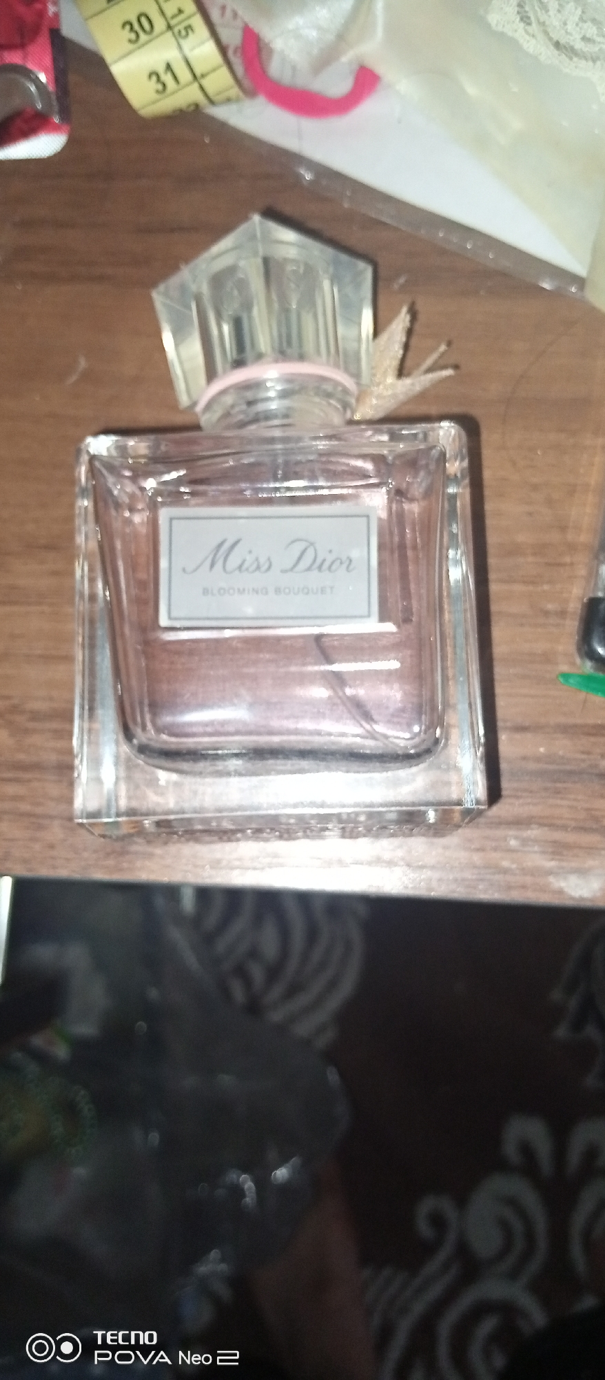 A close-up, eye-level, indoor shot displays a glass bottle of Miss Dior Blooming Bouquet perfume. The bottle is rectangular with a clear cap and a pink band around the neck. The label on the bottle reads "Miss Dior" in cursive and "BLOOMING BOUQUET" in smaller text. The bottle contains a pinkish liquid. The background consists of a wooden surface and other out-of-focus objects. The bottom of the image shows a blurred reflection of the surroundings, including the "TECNO POVA Neo 2" logo. The image is taken in a domestic setting, likely a home, within the city of Tahmach, Uzbekistan.