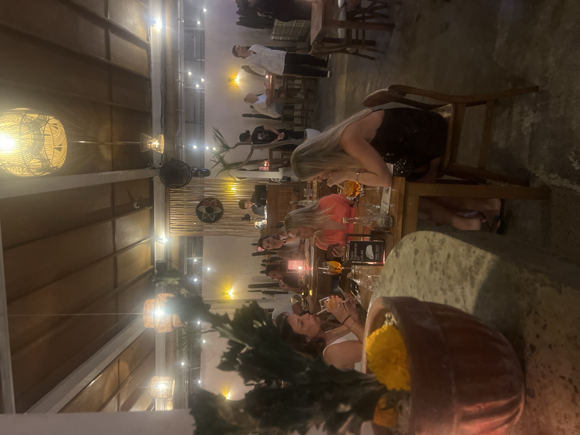 The image shows a dimly lit restaurant interior in North Kuta, Indonesia, with a rustic ambiance. Several patrons are seated at tables, some of whom are drinking from glasses with orange-colored beverages. In the foreground, a large terracotta pot holds a spiky plant with a yellow flower. The ceiling features woven pendant lights. In the background, a man in a white shirt stands near a table, and other individuals are visible seated and standing in the restaurant. A sign on a table reads "TACO TUESDAYS AT LOLAS FREE TACO". The overall setting suggests a lively dining establishment.