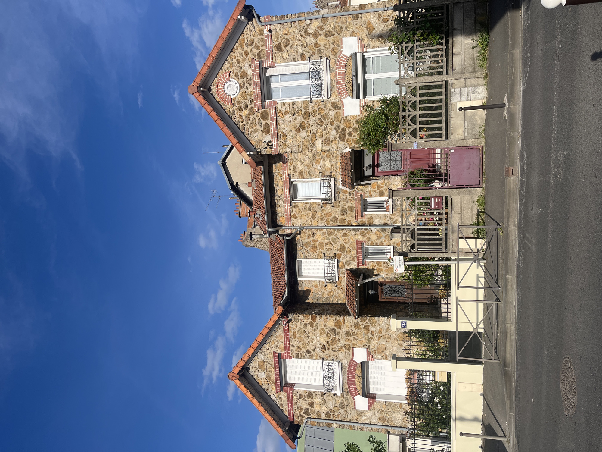 The image depicts a residential street scene in Vitry-sur-Seine, France, under a clear blue sky with scattered white clouds. The setting appears to be a sunny day, likely midday or early afternoon, given the bright illumination.
The main subjects are a row of stone and brick houses, closely situated along a paved street. The houses feature facades constructed from irregular, light-colored stones interspersed with reddish-brown brickwork. The bricks are used to outline windows, create decorative arch patterns above some windows, and form vertical accent strips. The roofs are covered with distinct reddish-orange tiles, and several white-framed windows with white louvered shutters are visible. Some windows also have elaborate black wrought-iron grates.
At street level, various metal fences and gates separate the properties from the sidewalk. One prominent house entrance features a dark red or maroon wooden door with decorative ironwork, beside which is a matching maroon metal gate. Another section of fencing is made of concrete or stone with decorative cut-outs. Green foliage and shrubs are visible behind some of the fences. The asphalt street runs parallel to the houses, featuring a manhole cover and a black bollard on the adjacent concrete sidewalk.
Notable text includes a white sign on a pole that reads "vitry en scene" and "point info" with a red logo. Further down the street, a house number "24B" is visible on a blue plaque attached to a white pillar. Another partially obscured street sign appears to indicate "Rue Gabriel Peri," which confirms the location. No people or active events are depicted in the static scene.