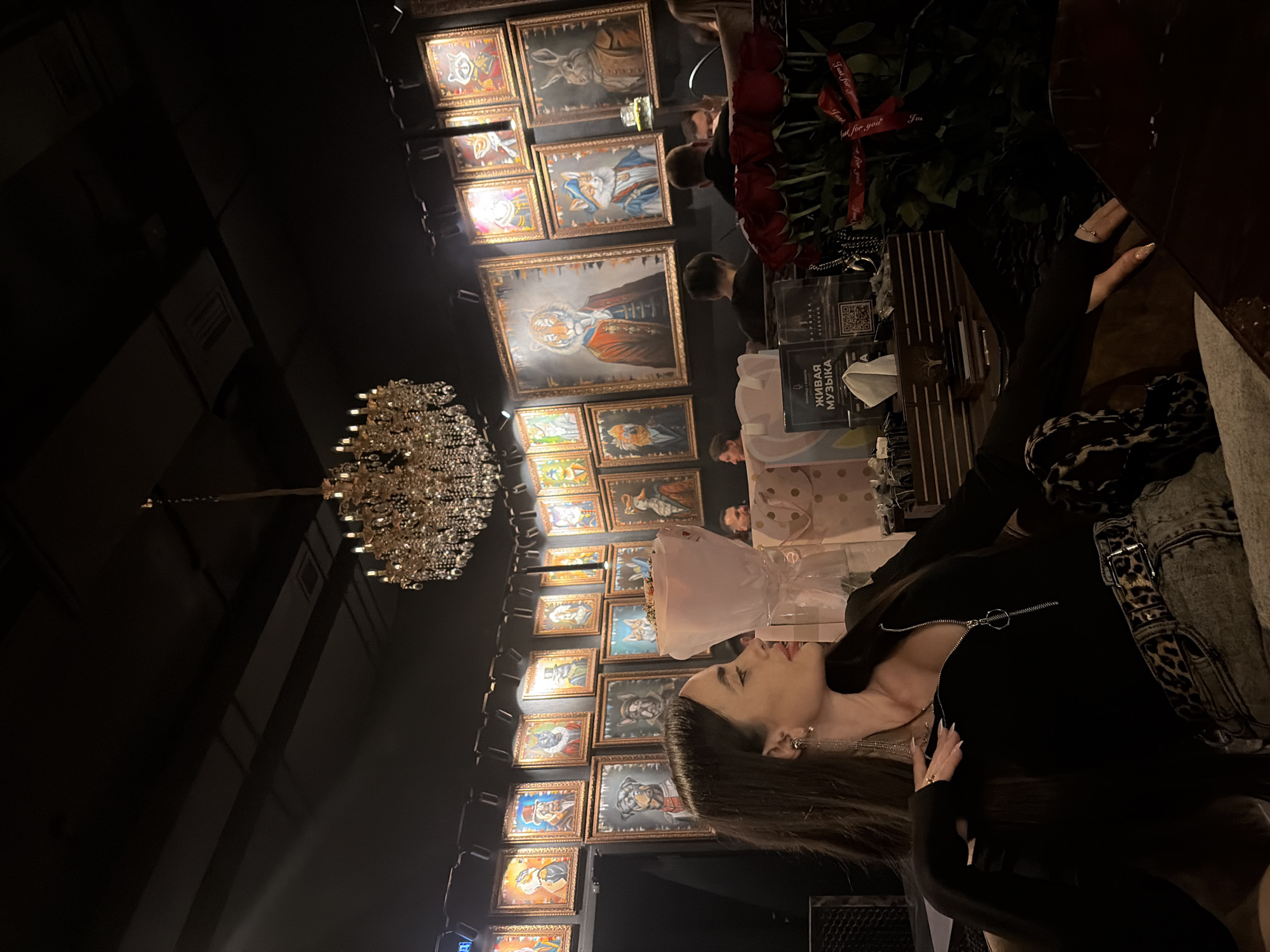 The image captures an interior scene. A dark-haired woman is visible, wearing a black zipped shirt. In the background, a wall is adorned with framed artwork. A chandelier hangs from the ceiling. The environment appears to be a restaurant. The restaurant is in Saint Petersburg, Russia, as identified by the menu card on the table written in the Russian language. Additionally, there are rose bouquets on the tables.