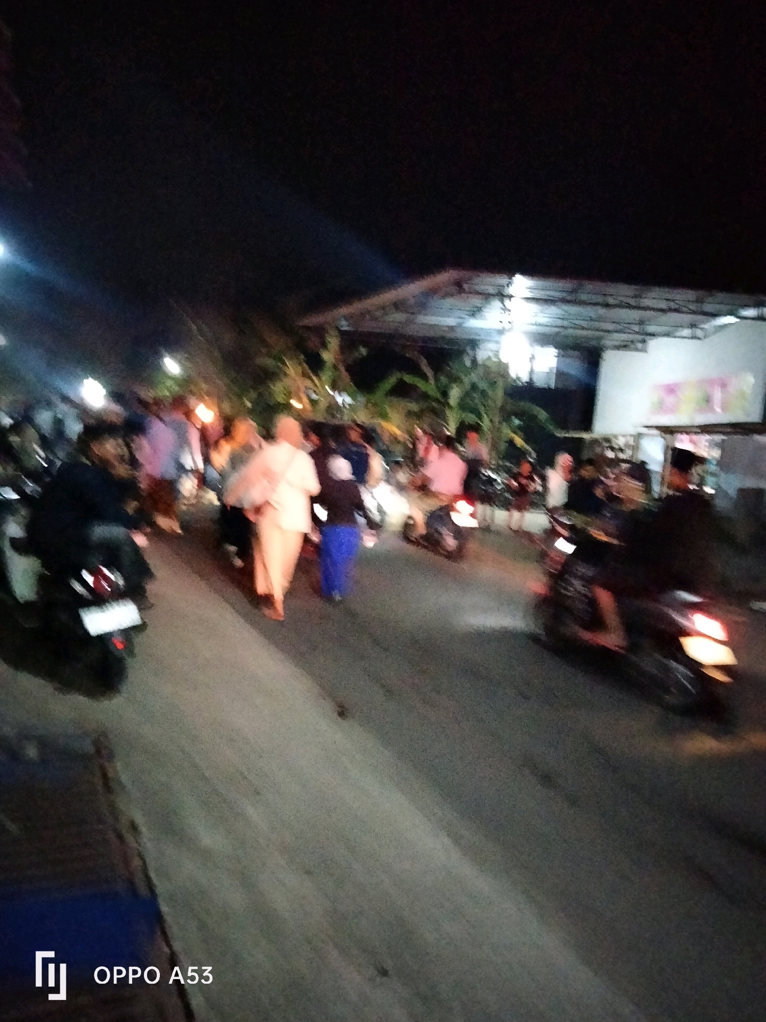 At night in Bendorejo, Indonesia, a street scene unfolds with several people on foot and on motorcycles. In the foreground, a person is seated on a dark-colored motorcycle, with its tail lights illuminated. Another motorcycle is in motion in the mid-ground on the right, its tail lights also lit. A group of individuals are walking on the side of the road. One person in the center, wearing a light-colored garment, appears to be carrying something. Another individual beside them is wearing a blue garment. In the background, a structure with a corrugated roof is visible, illuminated by lights. Trees are present on the left side of the image. The overall scene depicts a busy street at night, possibly during a local event or gathering.