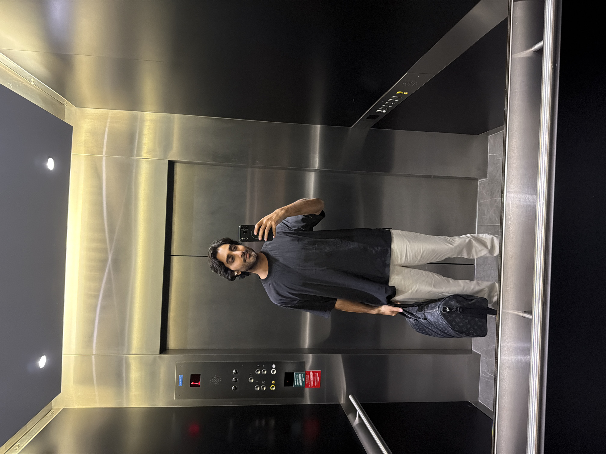 The image depicts a young man with dark hair and a beard, wearing a black t-shirt and off-white pants, standing inside an elevator and taking a selfie with his phone. He is holding a dark-colored duffel bag with a patterned design in his left hand. The elevator is metallic with bright lighting, and an elevator control panel with buttons and a digital display showing the number '1' is visible on the left wall. The floor of the elevator appears to be tiled in grey and white marble. The man is positioned in the center of the frame, looking directly at the camera. The elevator doors are closed, and the reflection of the man and the elevator interior is visible on the polished surfaces. There are no discernible landmarks or signs that would indicate the location is in London, United Kingdom.