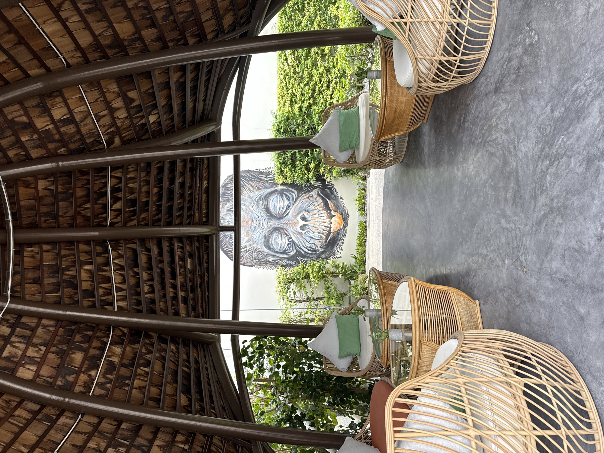The image shows an outdoor seating area within a thatched-roof pavilion in the Ko Samui District, Thailand. The pavilion's interior features a conical, wooden-slatted ceiling supported by dark brown structural beams. The floor is polished concrete. Several rattan armchairs with light-colored cushions and green accent pillows are arranged around glass-topped rattan coffee tables. Each coffee table has a small potted plant. In the background, through the open sides of the pavilion, lush green foliage and a large mural of a gorilla's face are visible on a white wall. The overall setting appears to be a relaxed, open-air lounge or waiting area.