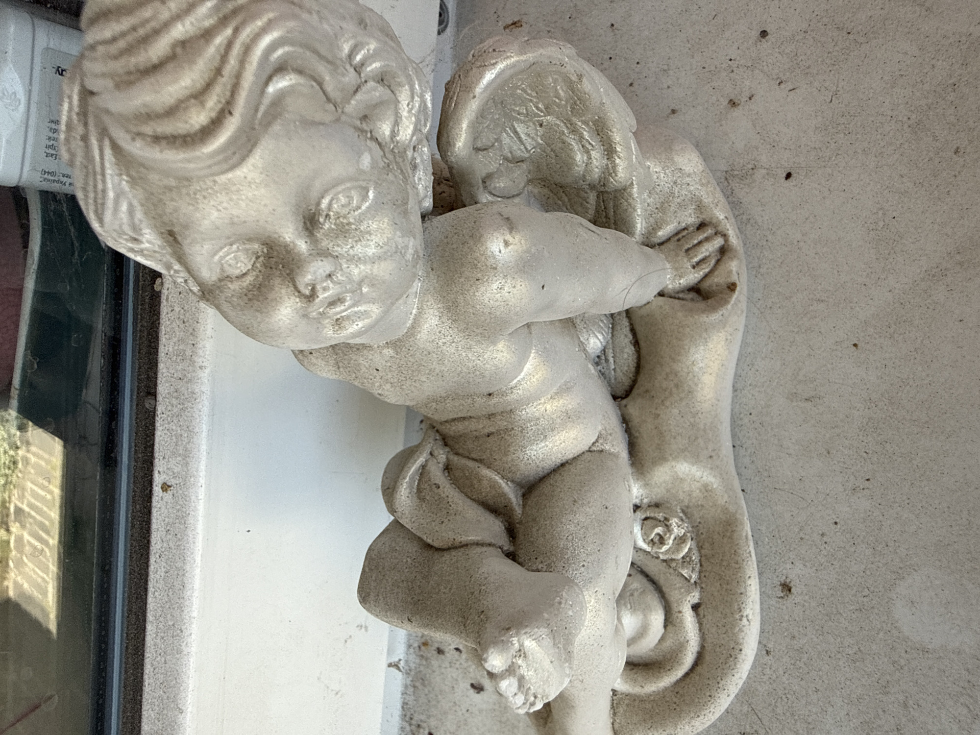 An aged, light-colored statue of a cherub reclines on a textured concrete surface. The statue, depicting a winged infant with detailed hair, is positioned diagonally across the frame. The cherub's head is turned to the left, with its eyes closed. One arm is bent, with a hand resting on a curved, elongated base. The other arm is extended downwards. The cherub's legs are crossed, with one foot visible. The statue appears weathered and has a slightly grainy texture. In the background, a dark windowsill with a black rubber seal is visible, above which is a window with a reflection of what appears to be a building or structure. The scene is located in Okhtyrka, Ukraine.