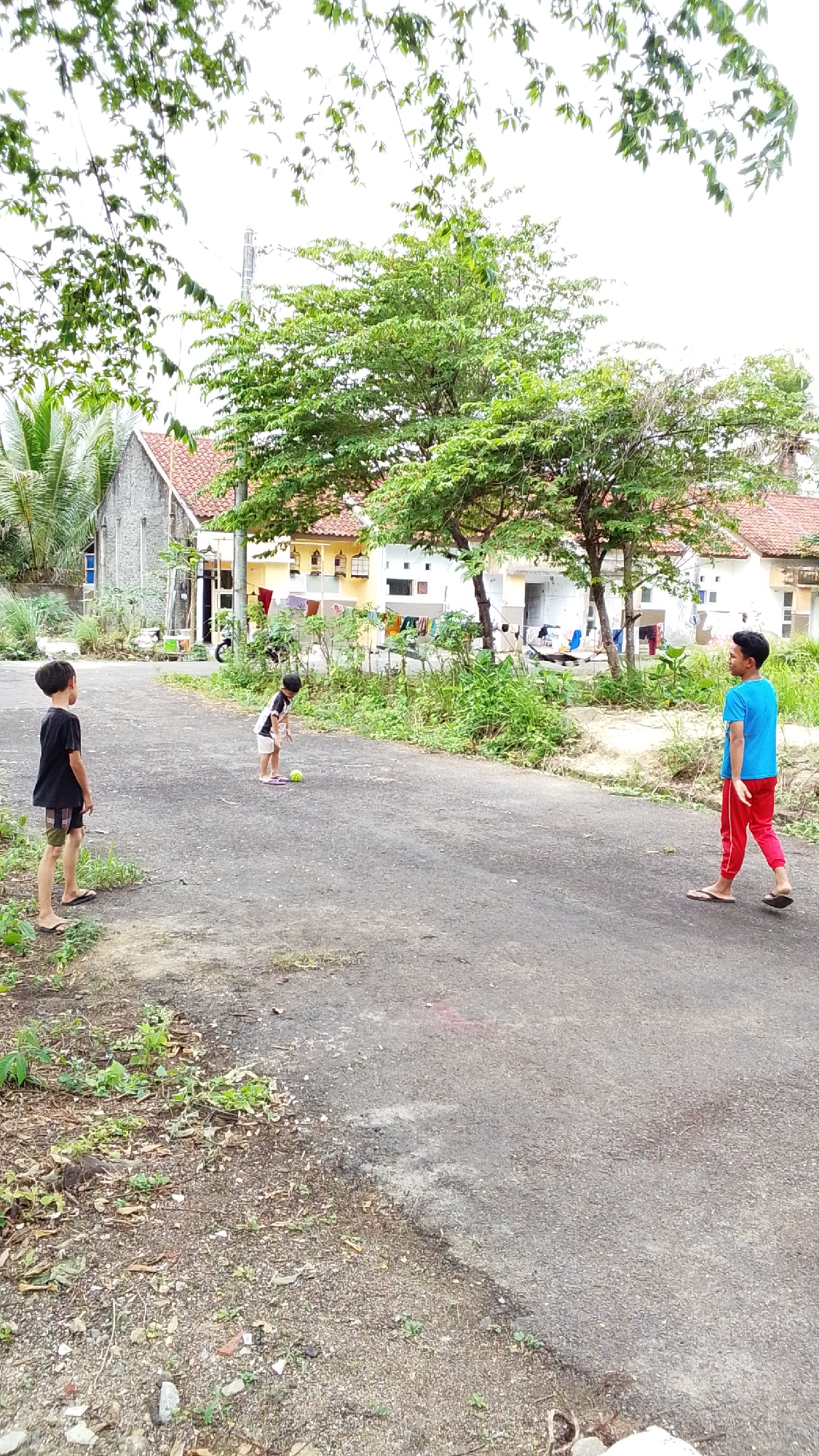 Three young boys are playing on a paved road in what appears to be a residential area. The boy on the left is facing away from the camera, wearing a black t-shirt and plaid shorts. In the center, a boy in a black and white striped shirt and white shorts is bent over, possibly interacting with a small, light-colored ball on the ground. To the right, a boy in a blue t-shirt and red pants is walking towards the center, looking back over his shoulder. The road is surrounded by vegetation and houses with red-tiled roofs are visible in the background. The sky is overcast. The scene suggests a casual outdoor activity in a village setting in Cintakarya, Indonesia.