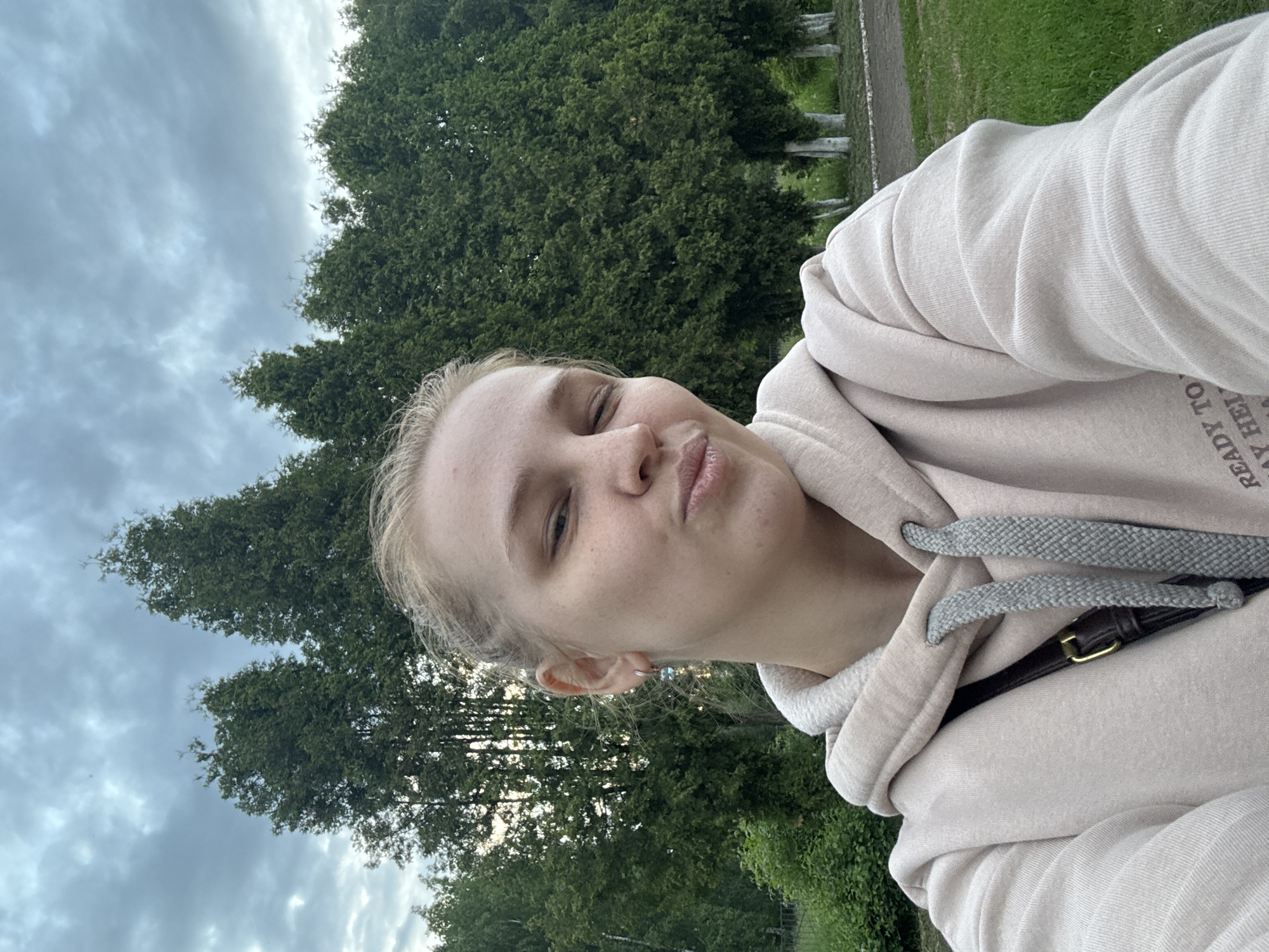 This is a close-up, angled selfie of a young woman in an outdoor setting in Rusanovo, Russia. The woman, with blonde hair, light skin, and freckles, is captured in a playful pose, squinting her right eye and pursing her lips. She is wearing a light beige or off-white hooded sweatshirt with gray, textured drawstrings. A dark strap with a small gold buckle, possibly from a bag, is visible across her chest, underneath the hoodie. A small, light-colored earring is seen in her left ear.
The background features dense green trees, suggesting a park or forested area. To her right, there is green grass adjacent to what appears to be a dirt or gravel path, with several short white posts or markings visible along its edge. The sky above is overcast with gray clouds, indicating cloudy weather during the daytime, possibly late afternoon or early evening. Part of the text "READY TO" and "AY HEI" is visible on the fabric of her hoodie, near her right shoulder. The overall mood conveyed is casual and lighthearted.