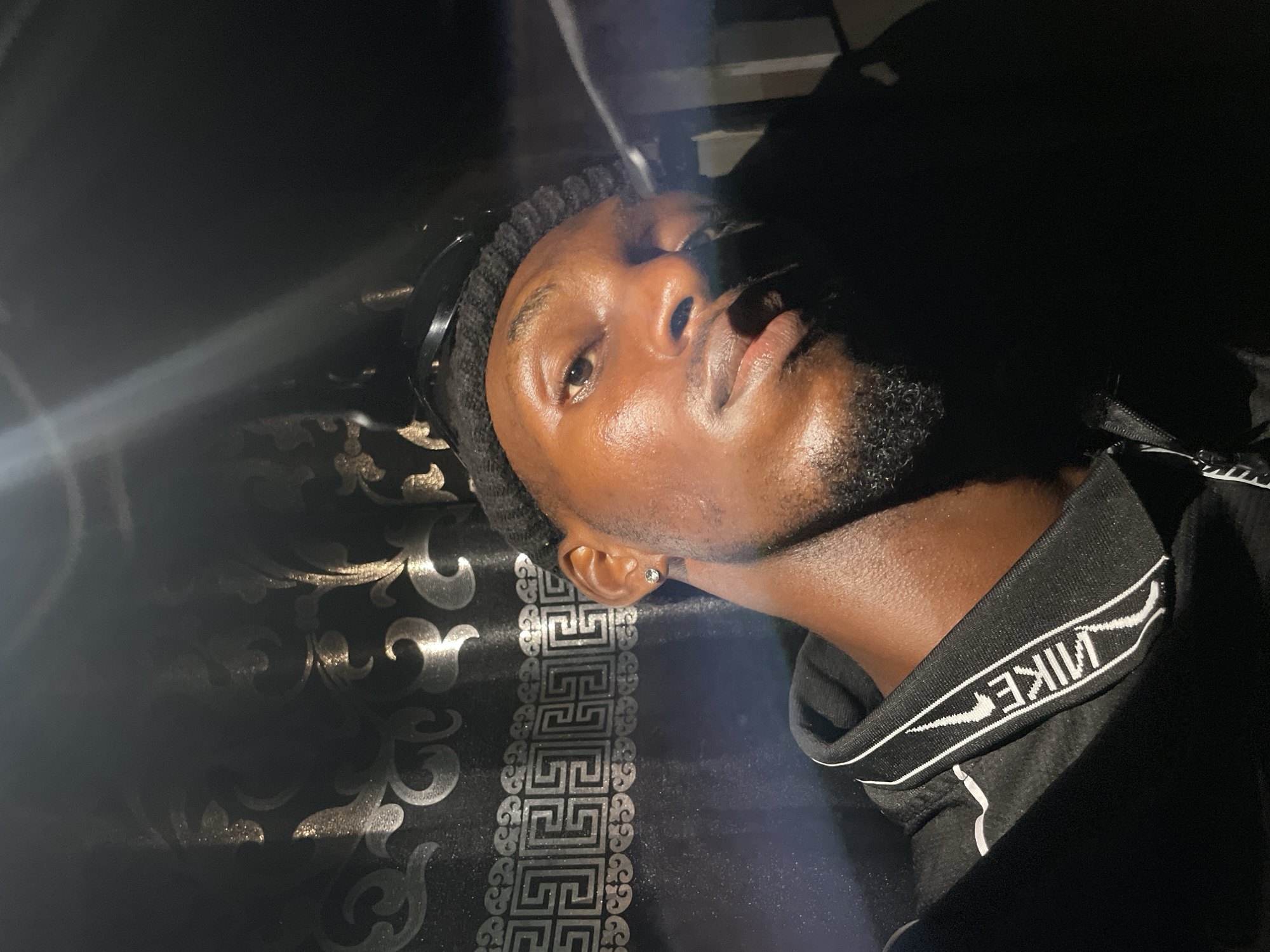 A close-up, angled shot depicts a dark-skinned individual with a trimmed beard, wearing a black beanie and a black Nike hooded sweatshirt. The background features a dark, patterned fabric with silver and gold accents. The location is unknown and cannot be determined from the image.