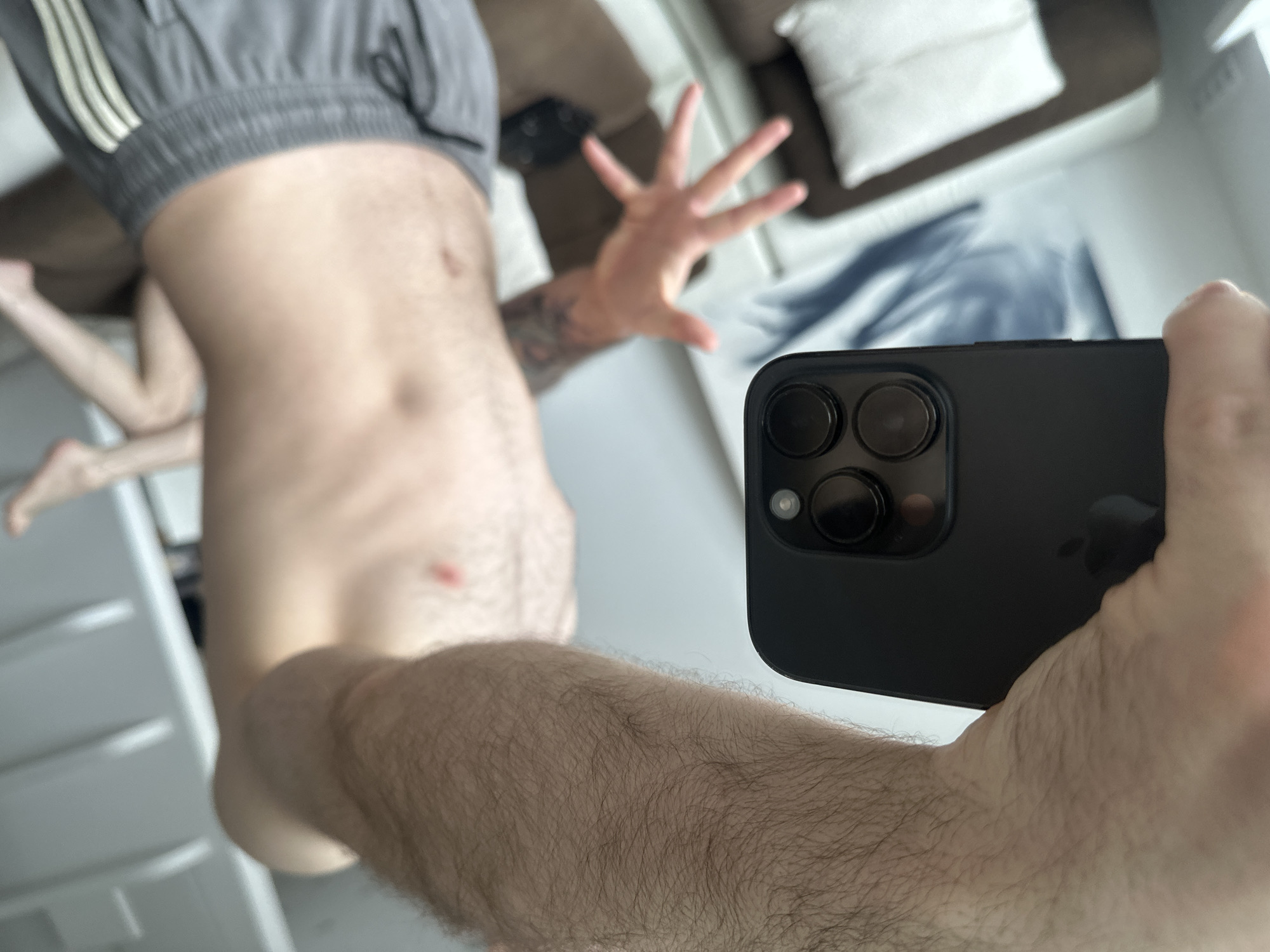A partially nude man in grey shorts is taking a selfie with a black iPhone. The image includes a view of his torso, legs, one arm holding the phone, and another arm extended. Background elements include a couch, pillows, and a white surface, potentially a mirror or a window. The location is not discernible in the image.