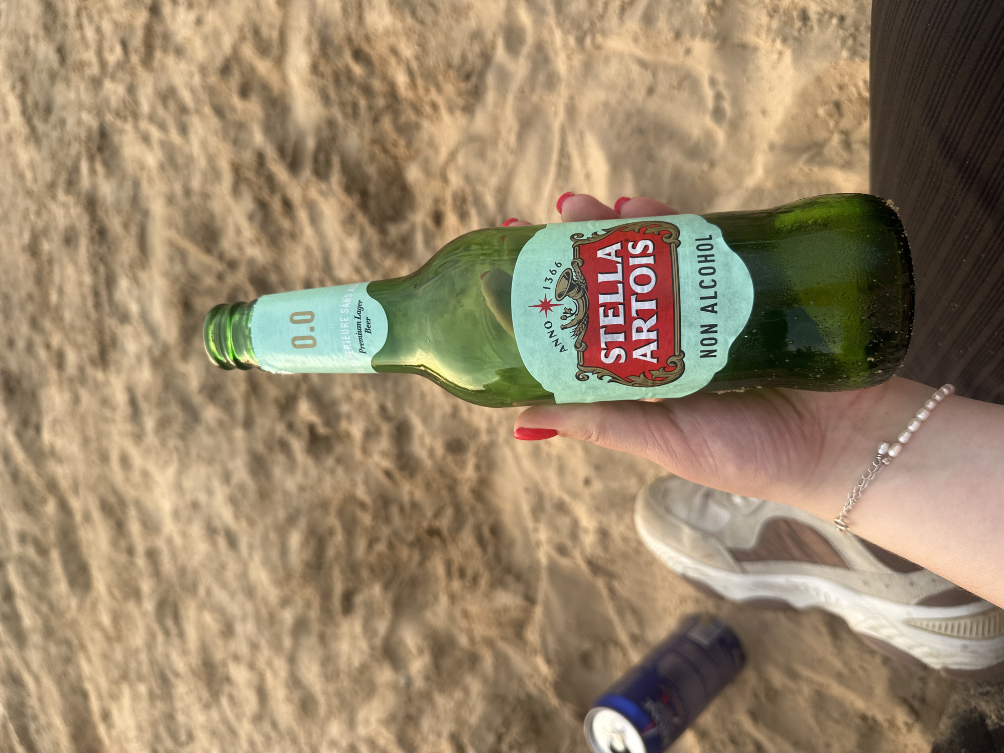 A person is holding a green glass bottle of Stella Artois Non Alcohol beer on a sandy beach. The bottle has a light blue label with red and white text and a crest. A blue soda can and a white athletic shoe are visible on the sand in the background. The person is wearing a bracelet with pearls. The image is taken in Lytkarino, Russia.