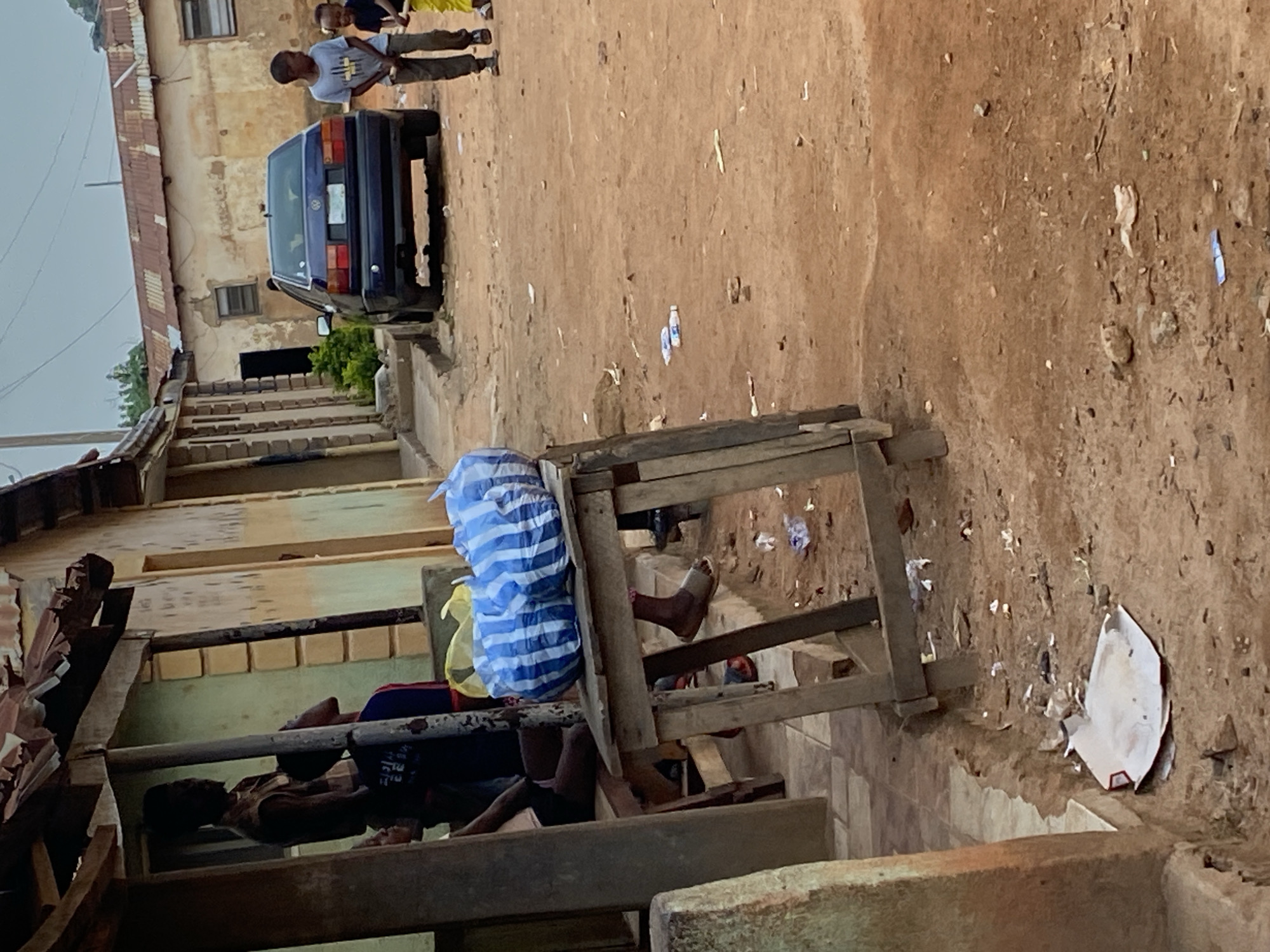 A street scene in Bukuru, Nigeria, shows a dusty, unpaved road in front of a row of weathered buildings. The buildings have corrugated metal roofs and faded paint. In the foreground, a wooden table is laden with two large blue and white striped plastic bags. A person's legs are visible underneath the table, wearing sandals. To the left, a man stands under an overhang, partially silhouetted. Further down the street, a blue Volkswagen car is parked. Two young boys are walking towards the viewer; the one on the left is wearing a light-colored t-shirt and dark pants, while the one on the right is partially obscured. The ground is littered with debris, including plastic bottles and wrappers. The overall impression is one of a somewhat disheveled but active urban environment.