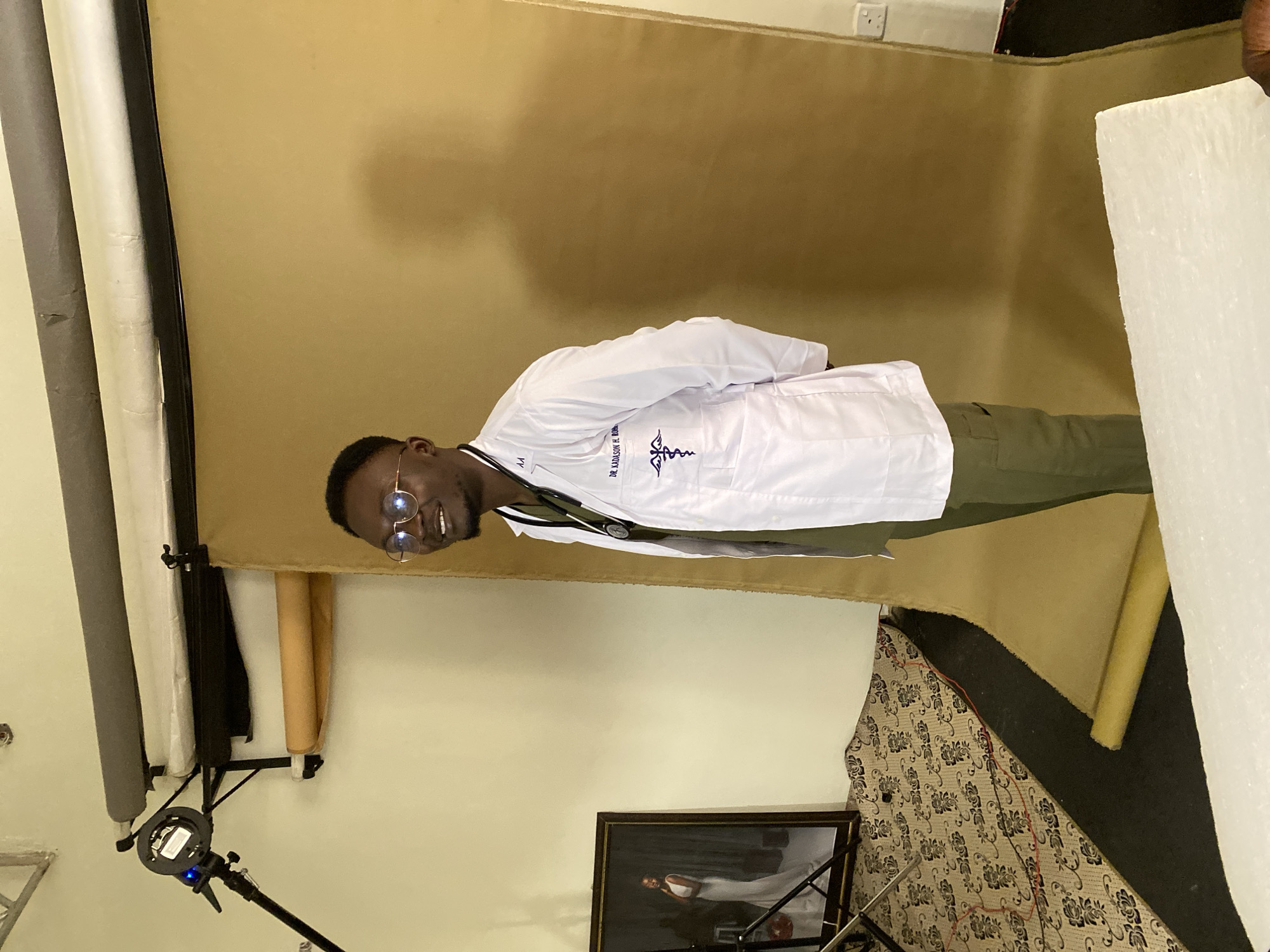 The image depicts a man wearing a white lab coat and glasses, standing in what appears to be a photography studio. The background consists of a large, tan-colored backdrop. To the left, there is a framed portrait of a woman in a white dress and a lighting setup with a stand and a light. A patterned rug covers the floor in the lower left portion of the image, and a black mat is visible beneath it. The man's lab coat has embroidery that includes a medical symbol and text that reads "DR. KADASON H. BOM." The overall scene suggests a professional portrait session is in progress, possibly for a medical professional. The image does not provide any explicit details about the location in Jos, Nigeria.