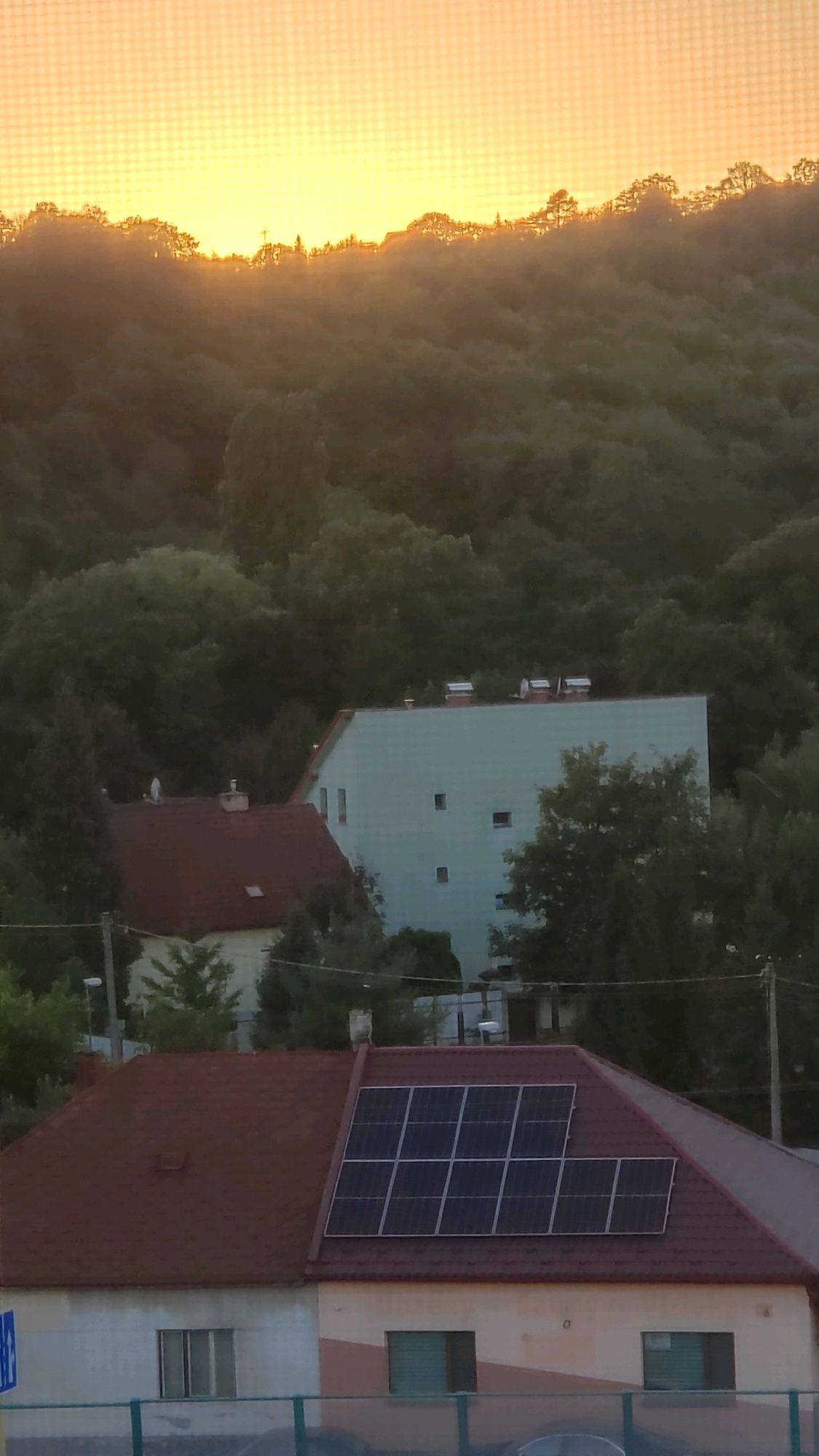The image shows a sunset over a residential area in Prešov, Slovakia. The sky is a warm orange and yellow, with a hazy glow. In the foreground, there are several houses with red roofs. One house has solar panels installed on its roof. Behind the houses, there is a densely wooded hill. The scene is peaceful and serene.