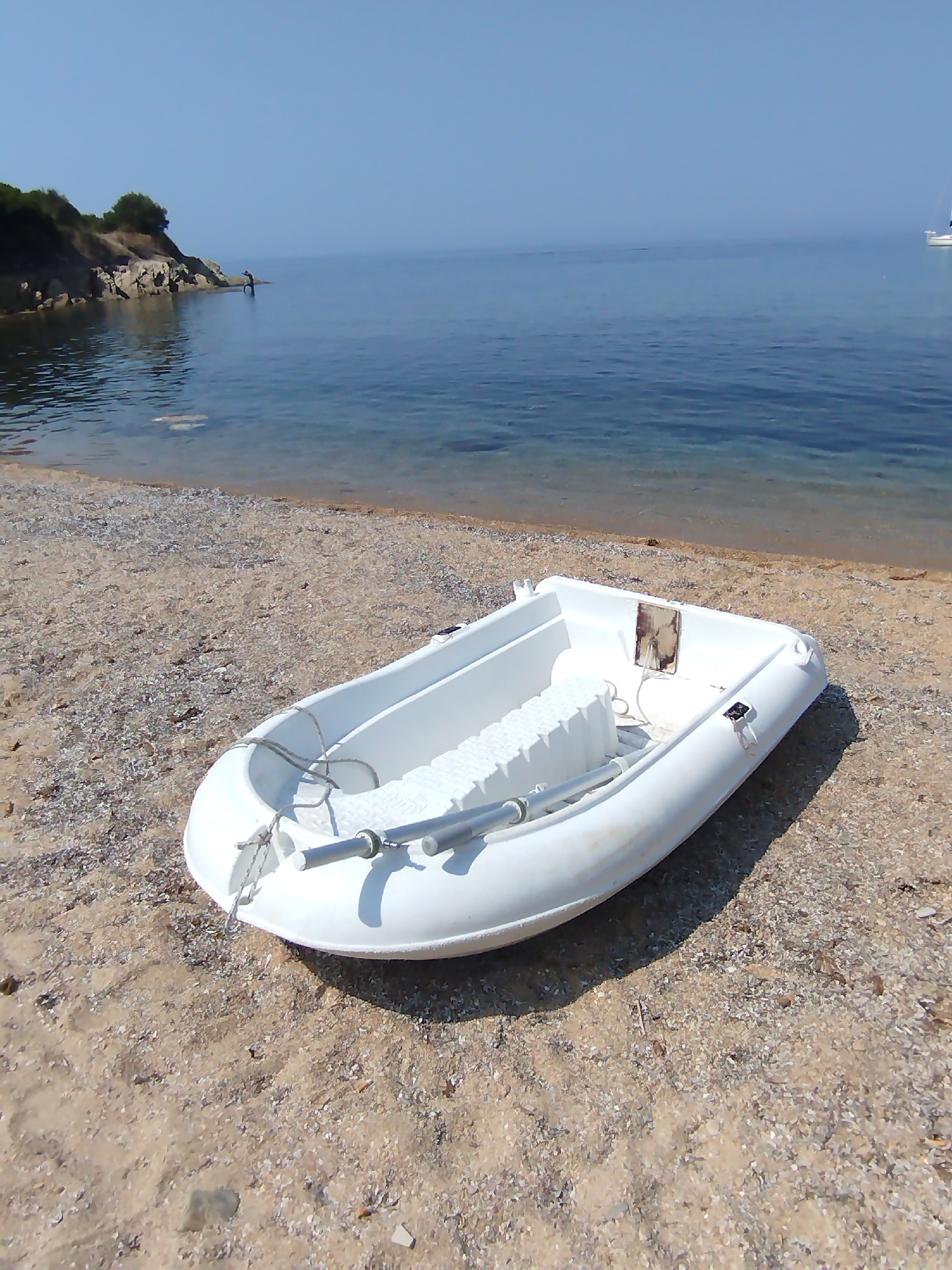 A white inflatable boat with silver oars lies on a sandy and pebbly beach. The boat is positioned in the foreground, angled towards the right side of the frame. Inside the boat, a row of white foam seat cushions is visible. The water is calm and blue, with a clear transition from shallow to deeper water. In the background, a rocky, tree-covered coastline extends to the left, with a person standing in the shallow water near the shore. A sailboat is visible in the far distance on the right horizon. The sky is clear and blue. The scene appears to be a tranquil coastal setting in Sithonia Municipal Unit, Greece.