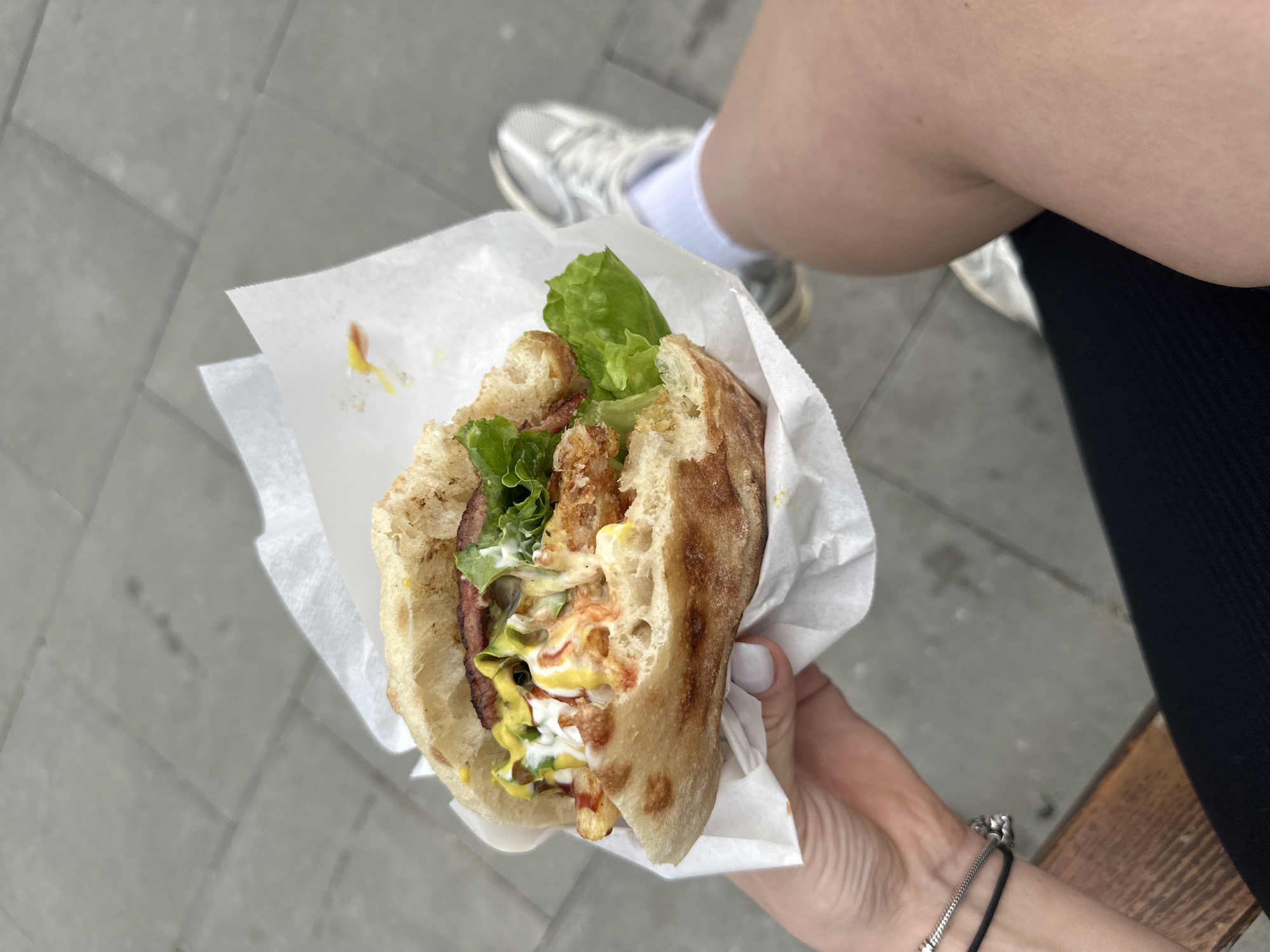 A person holds a partially eaten sandwich in a white paper wrap. The sandwich appears to contain lettuce, some kind of fried ingredient (possibly chicken or fish), bacon, and several types of sauces, including mustard and a white creamy sauce. The bread is crusty and light in color.
The person holding the sandwich has pale skin and painted nails. They wear bracelets on their left wrist. The person is seated, wearing black pants and a white sock and a grey sneaker on their left foot.
The background is a grey tiled surface. The image was taken in daylight. The location is unknown and cannot be identified as the city of Plovdiv, Bulgaria based on the provided visual information.