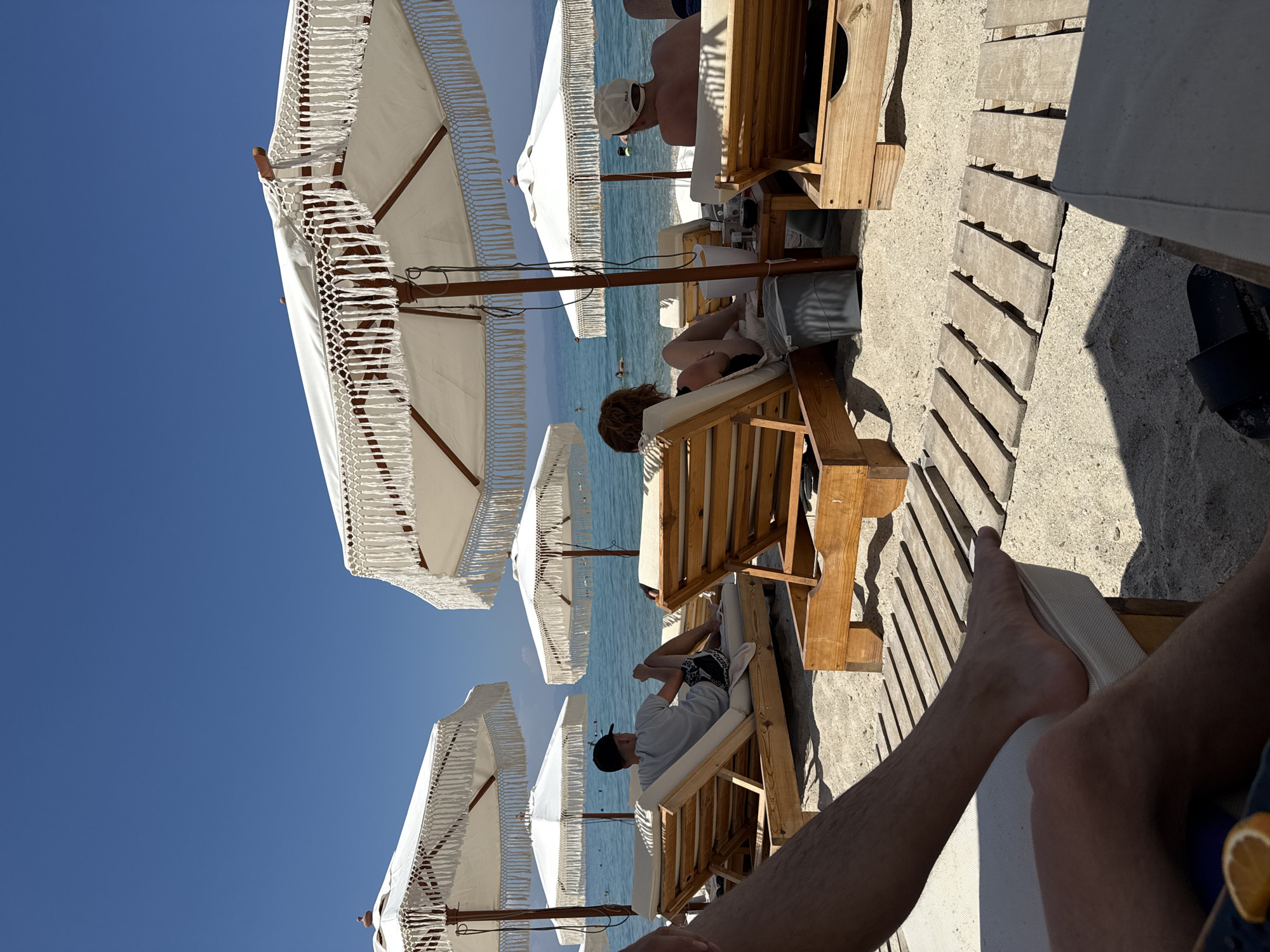 The image captures a sunny day at a beach in Pallini Municipal Unit, Greece. In the foreground, a person's legs and feet are visible, resting on a white padded lounge chair. The sand is a light beige color, and wooden planks are laid out on the sand in a pathway.
Further back, several white beach umbrellas with decorative fringe provide shade for wooden lounge chairs. In the middle ground, a person wearing a black cap and a light-colored t-shirt is seated on a lounge chair, facing the sea. Their legs are crossed, and they appear relaxed. Beside their chair is a wooden table with a drink container and other items.
Another person with reddish-brown hair is seated on a lounge chair under a different umbrella, facing away from the camera. To the right, a man wearing a white cap is also seated on a lounge chair, facing the water. The sea is a clear blue, with several people swimming and bathing in the distance. The sky is a vibrant, cloudless blue, indicating a warm and clear day. The overall scene depicts a relaxed atmosphere at a beach resort.