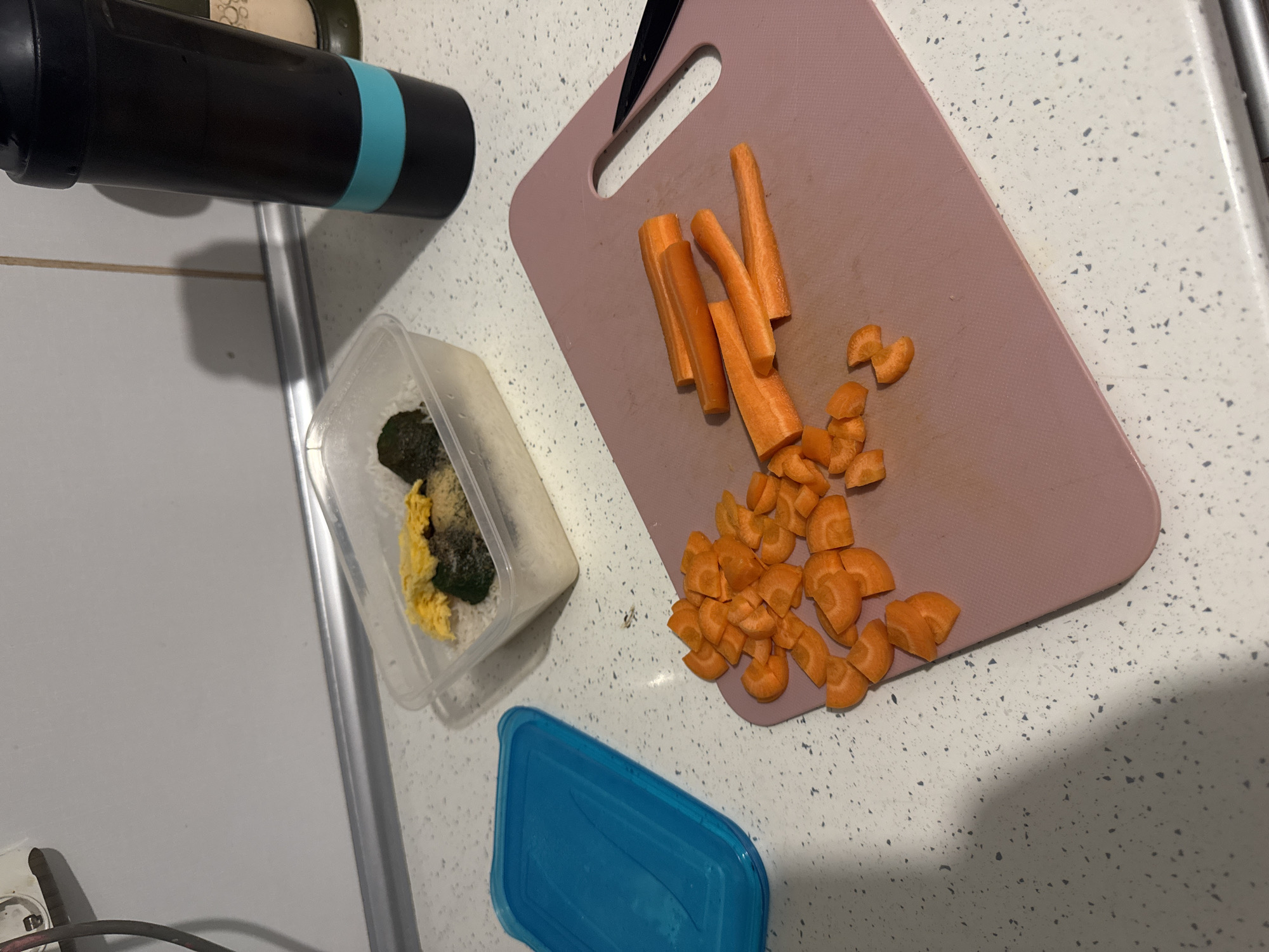 On a countertop with a speckled white surface, a pink cutting board holds partially sliced carrots. A container to the left contains food items, and a blue lid sits next to it. A black object with a blue band is positioned in the upper left corner.
