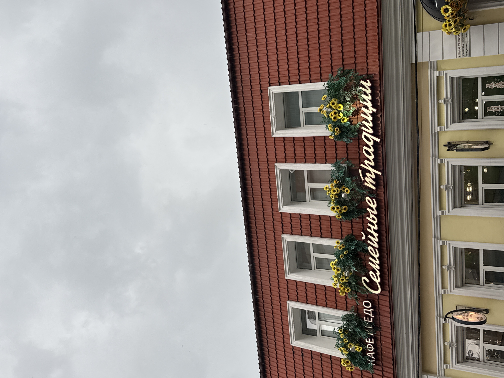 The image shows the facade of a building in a city, under a cloudy sky. The building has a red tiled roof with four white-framed windows. Below each window, there are flower boxes filled with sunflowers and greenery. A neon sign above the windows reads "КАФЕ НЕДО Семейные традиции". Below the red roof, the building's facade is painted yellow, with more white-framed windows and decorative elements. There are also signs visible on the yellow facade, including one with a WiFi symbol. The overall impression is of a charming building in an urban setting, possibly a cafe.
