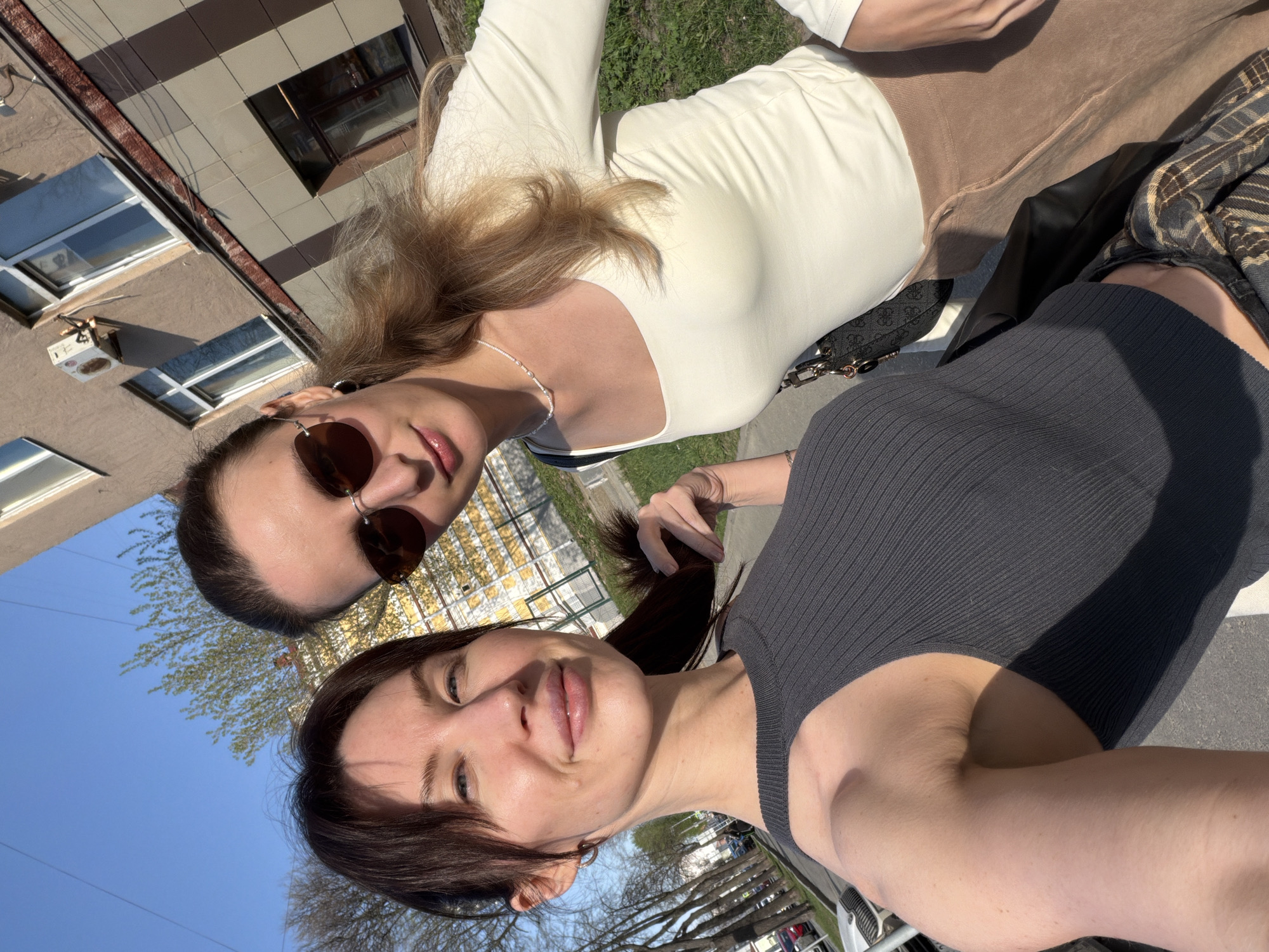 Two women are posing for a selfie outdoors. The woman on the top, with blonde hair, is wearing sunglasses, a white top, and a pearl necklace. The woman on the bottom, with dark hair pulled back in a ponytail, is wearing a gray top. Buildings, trees, and blue sky are visible in the background. The location is in the city of Perm, Russia.