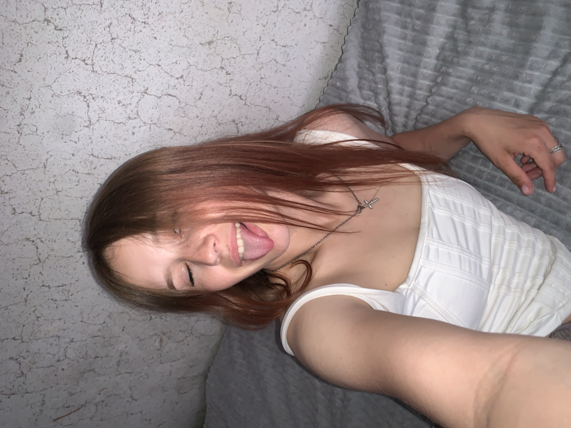 A young woman with auburn hair is lying on a gray textured surface, possibly a bed or couch, with a white textured top. She is sticking her tongue out with her eyes closed and appears to be taking a selfie. She is wearing a silver necklace with a cross pendant. The background includes a wall with a cracked or textured finish. The scene is indoors. There is no evidence to indicate it is located in the city of Фоки, Russia.
