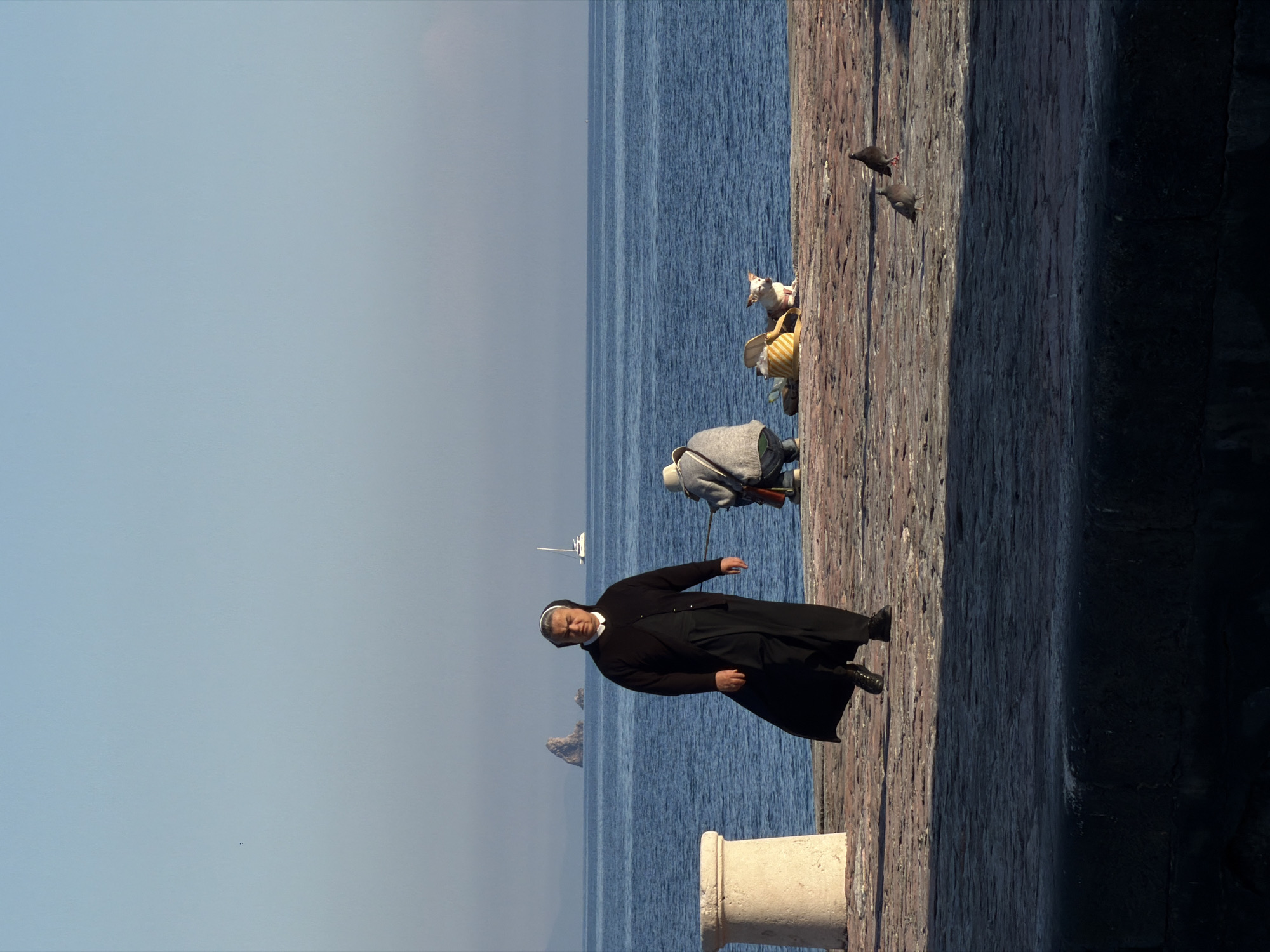 The image displays a bright, daytime scene on a seaside promenade in Petrovac centar, Montenegro. The view is along a paved walkway bordered by a dark, textured wall on the right and the deep blue sea on the left.
In the foreground, a woman wearing a black nun's habit, including a white wimple and veil, walks towards the viewer with a composed expression. A light-colored stone pillar or bollard is partially visible to her right. Further along the promenade, another person, dressed in a light-colored hooded garment and trousers, is seen from behind, seemingly bending or looking down at something. Nearby, a small white dog with distinctive markings is present, possibly in a carrier or being held. Two dark birds, likely pigeons, are also on the promenade surface in the mid-ground.
The expansive sea stretches to the horizon, exhibiting subtle ripples under a clear, light blue sky. In the mid-distance on the water, a white sailboat is visible. Beyond it, two rocky islets or small landmasses emerge from the sea.
The scene conveys a sense of tranquility and leisure. The clear sky and bright lighting indicate good weather, likely during the mid-morning or afternoon. There is no visible text in the image.