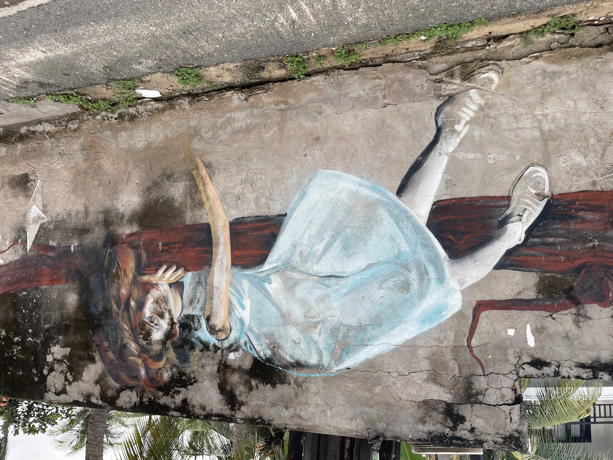 A mural of a girl in a light blue dress, painted on a crumbling concrete wall. The wall is cracked and stained, with some vegetation growing along the top edge near the pavement. The girl is depicted hanging upside down, with her shoes and legs pointing upwards. Her red hair falls around her face. The mural appears to be painted on an exterior wall in Phu Thuy Ward, Vietnam. Part of the wall extends into the distance near trees.