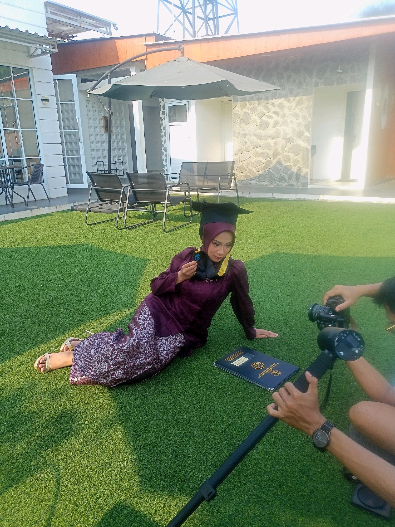 A graduation photoshoot is taking place outdoors on a bright, sunny day in Blambangan, Indonesia. A woman, dressed in a purple graduation gown and cap, is posing on a patch of green artificial turf. She is lying on her side, with her left arm extended and her right hand holding a medal. In front of her are two blue graduation diplomas. Two photographers are actively capturing the scene, with one operating a professional camera and the other holding a lighting device. In the background, a building with stone accents and outdoor seating furniture is visible. The scene conveys a celebratory and professional atmosphere.