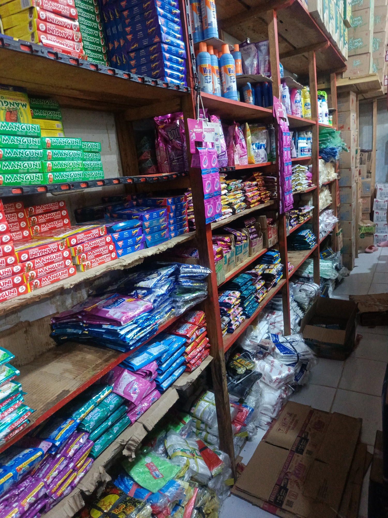 The image depicts the interior of a small retail store, likely a *warung* or convenience store, located in Ilotidea, Indonesia. The store is packed with shelves filled with various household goods and consumer products, predominantly packaged items. On the shelves, there are stacks of brightly colored boxes and bags, including what appear to be laundry detergent packets, cleaning supplies, and possibly food items. Several shelves are dedicated to stacked boxes of what seems to be mosquito repellent coils, judging by the packaging and the brand name "MANGUNI". Other visible items include bottles of cleaning liquids, sachets of powdered goods, and plastic bags containing more merchandise. The store appears somewhat cluttered, with items also stacked on the floor in some areas, suggesting a busy retail environment. The lighting is artificial, contributing to a somewhat dim ambiance. No people are visible in the image, but the presence of goods and shelving indicates a commercial space. The flooring is tiled.