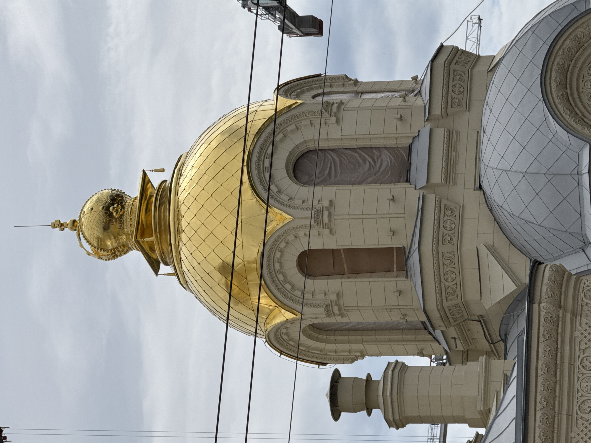 The image shows a close-up of the golden dome of a church in Saint Petersburg, Russia. The dome is topped with a cross and ornate detailing. The church has a light-colored facade with arched windows. There are visible construction elements, including cranes and scaffolding, suggesting renovation or construction work is underway. Power lines are visible in the foreground, crisscrossing the frame. The sky is overcast. The specific location within Saint Petersburg is not identifiable from the image alone.