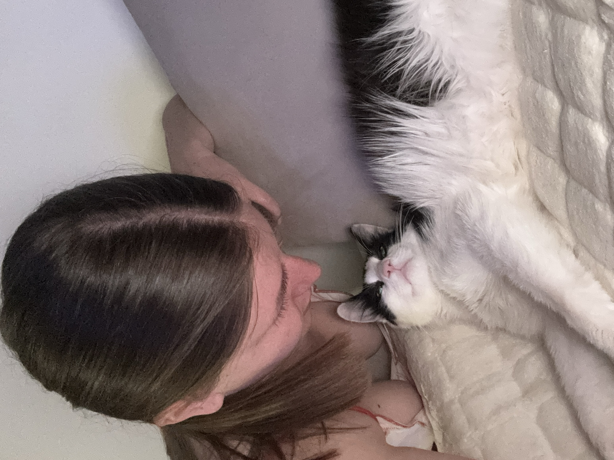 A woman lies on a bed next to a black and white cat. The woman has dark hair parted in the middle. Her face is turned towards the cat. The cat lies on its back with its face turned upwards. The bed is covered with a white quilted blanket. A gray pillow is positioned behind the woman's head. The location cannot be determined.