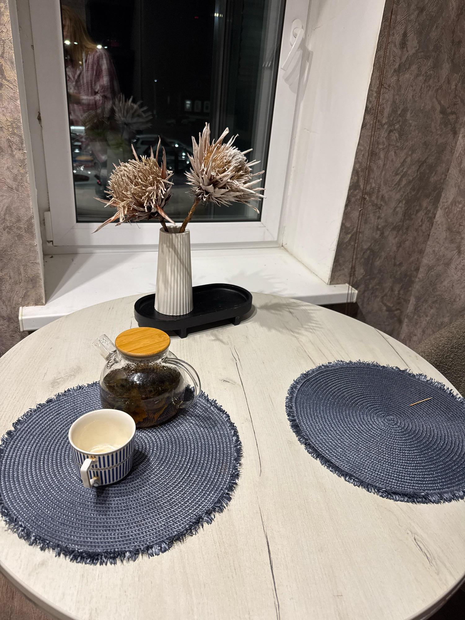 An indoor shot captures a simple, domestic scene on a light-colored, wood-grain patterned round table, likely in Valday, Russia. The table is set with two dark blue, oval woven placemats, each with a fringed edge. One placemat holds a clear glass teapot filled with dark tea and a wooden lid, accompanied by a white and blue striped teacup. The second placemat is empty except for a single toothpick. Behind the table, a white windowsill frames a view of a nighttime outdoor scene, with reflections of a person in pink pajamas visible in the window glass. On the windowsill, a fluted white vase holds two dried, spiky decorative plants. A black oval tray rests behind the vase. The background features textured, dark brown curtains.