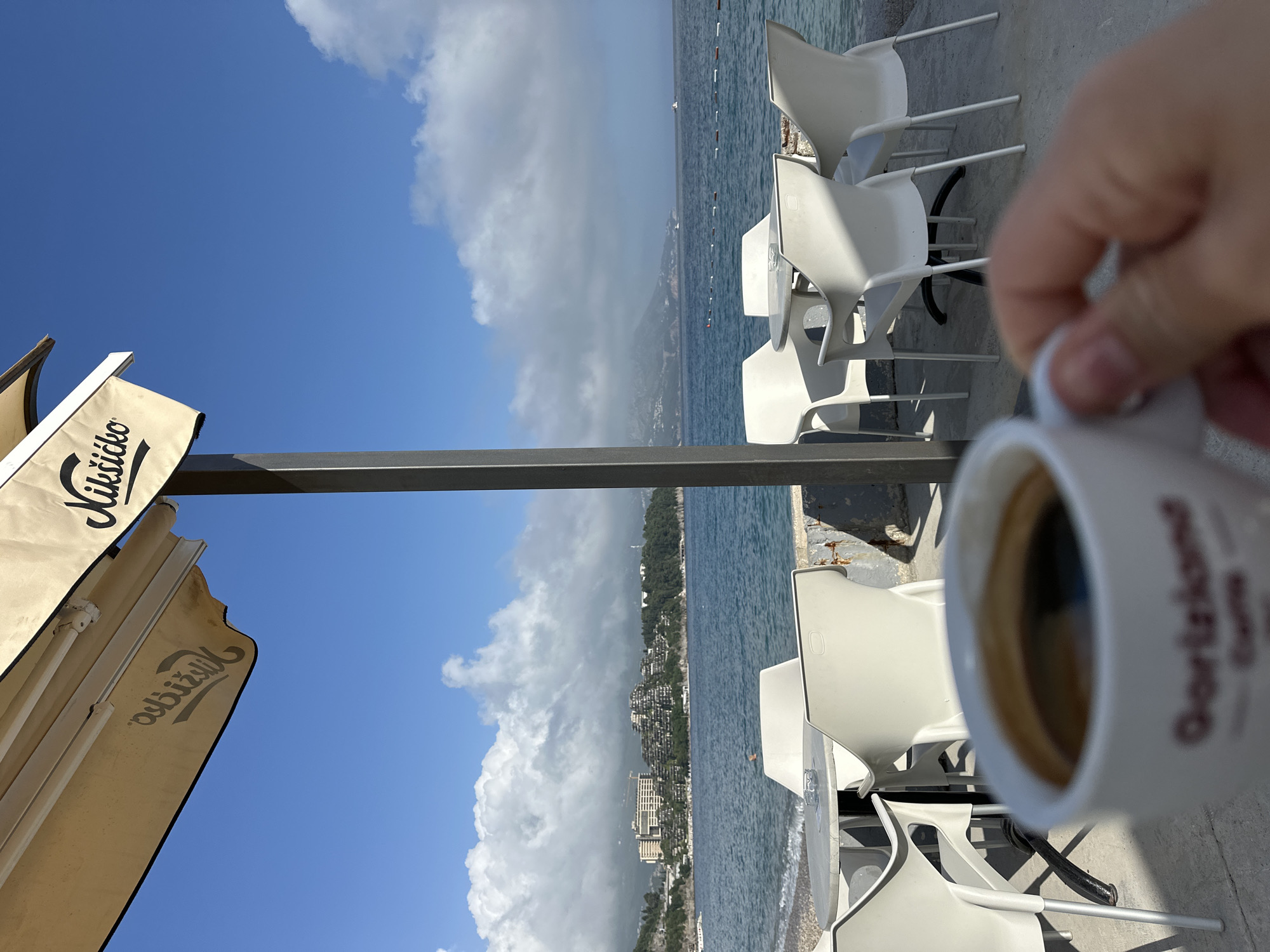 The photograph depicts an outdoor café scene in Budva, Montenegro. The foreground features a hand holding a cup of coffee, obscuring some of the background. Behind the coffee cup are several stacked white plastic chairs and tables, typical of outdoor café furniture. A metal beam runs horizontally across the middle of the image, partially blocking the view. Beyond the chairs and beam, the Adriatic Sea stretches towards a coastline lined with buildings. The sky is blue with scattered clouds, creating a contrast between the bright sky and the darker sea. To the left, the corner of a beige-colored awning is visible, displaying the brand name "Nikšićko" in Cyrillic script. The orientation of the photograph is horizontal, but the original image might have been taken vertically. The focus seems to be on the coffee cup, blurring the background details slightly.