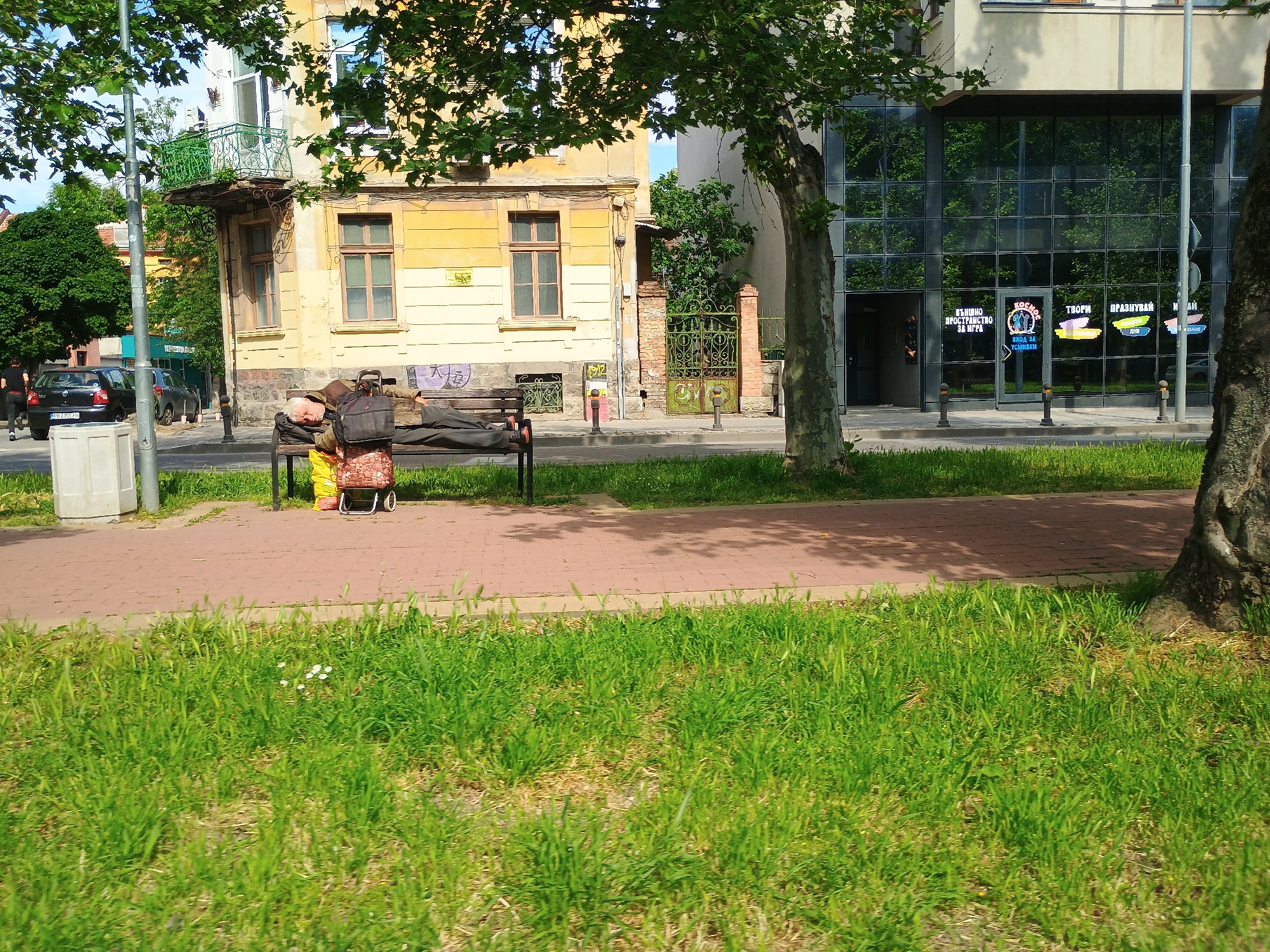 A man is lying on a park bench in front of a yellow building with a green balcony in the city of Unknown, Unknown. Next to the bench is a rolling cart and several bags. Across the street is a modern building with large glass windows and signage. The foreground is grassy. The scene is illuminated by daylight.