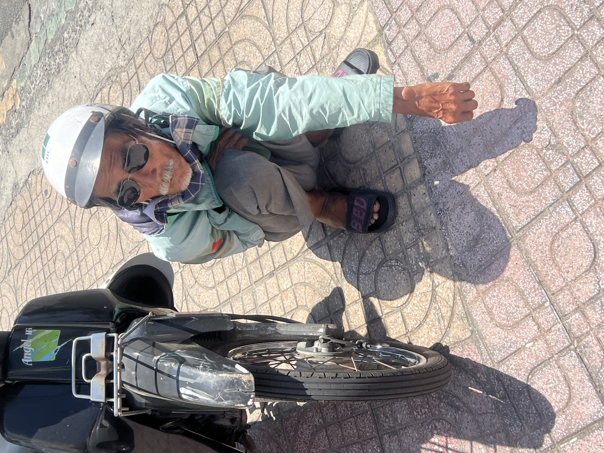 The image is a high-angle shot of a man next to a motorbike on a tiled sidewalk. The man is wearing a white helmet, sunglasses, a light green jacket, and gray pants. He's crouching next to the front wheel of the black motorbike. The motorbike has a sticker that reads "Angel Hi". The location appears to be a sidewalk in a built environment. The ground is made of square tiles with a speckled pattern of pink and beige. Based on contextual clues, the location is Nha Trang Ward, Vietnam.