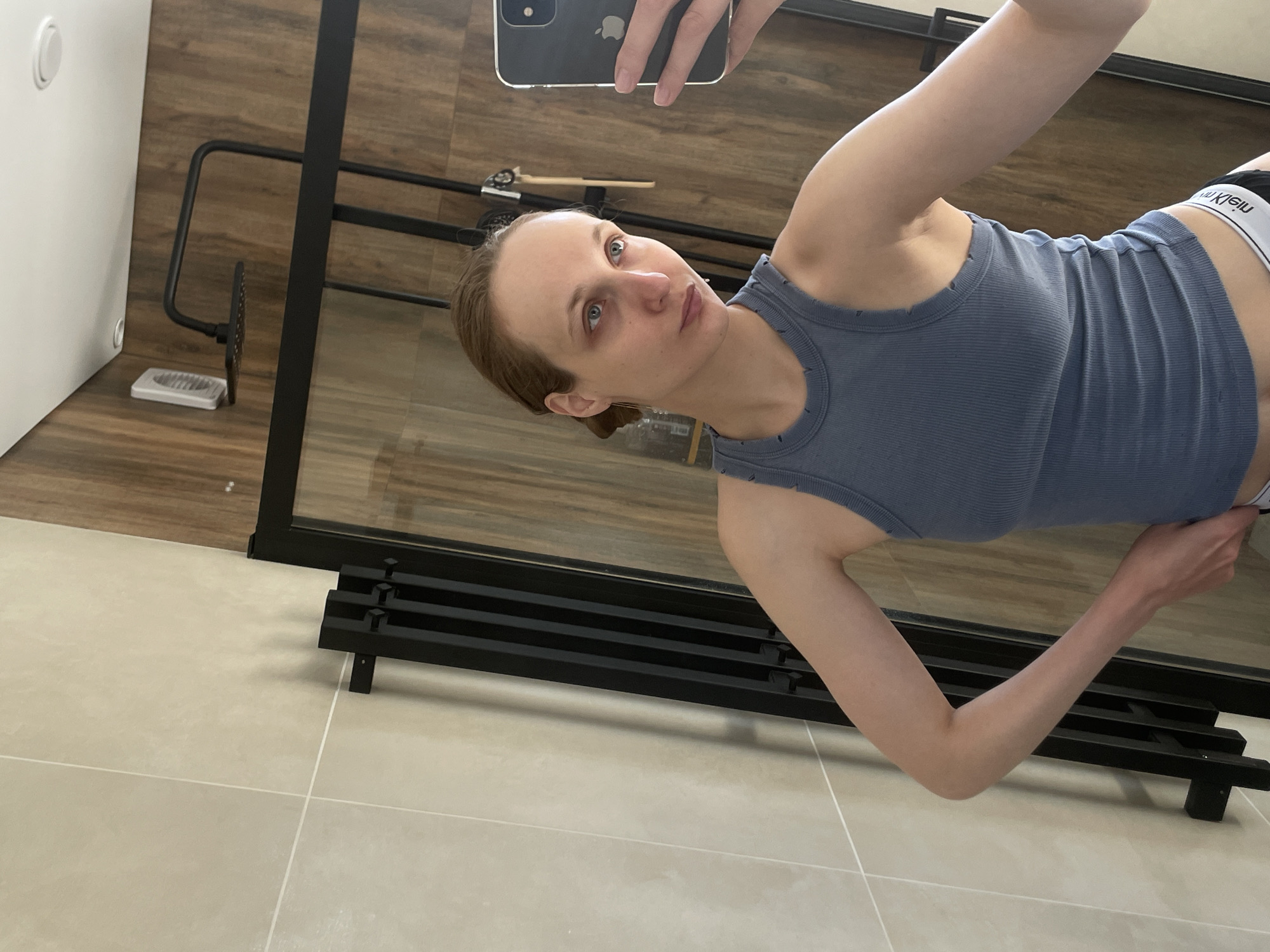 A woman with short brown hair and blue eyes stands in a bathroom in Подшивалово, Russia, taking a selfie. She is wearing a blue tank top and white underwear with a black waistband. The bathroom has beige tile flooring and wood-look wall paneling. A black metal towel rack is visible in the background. The woman holds a black smartphone up to take the picture.